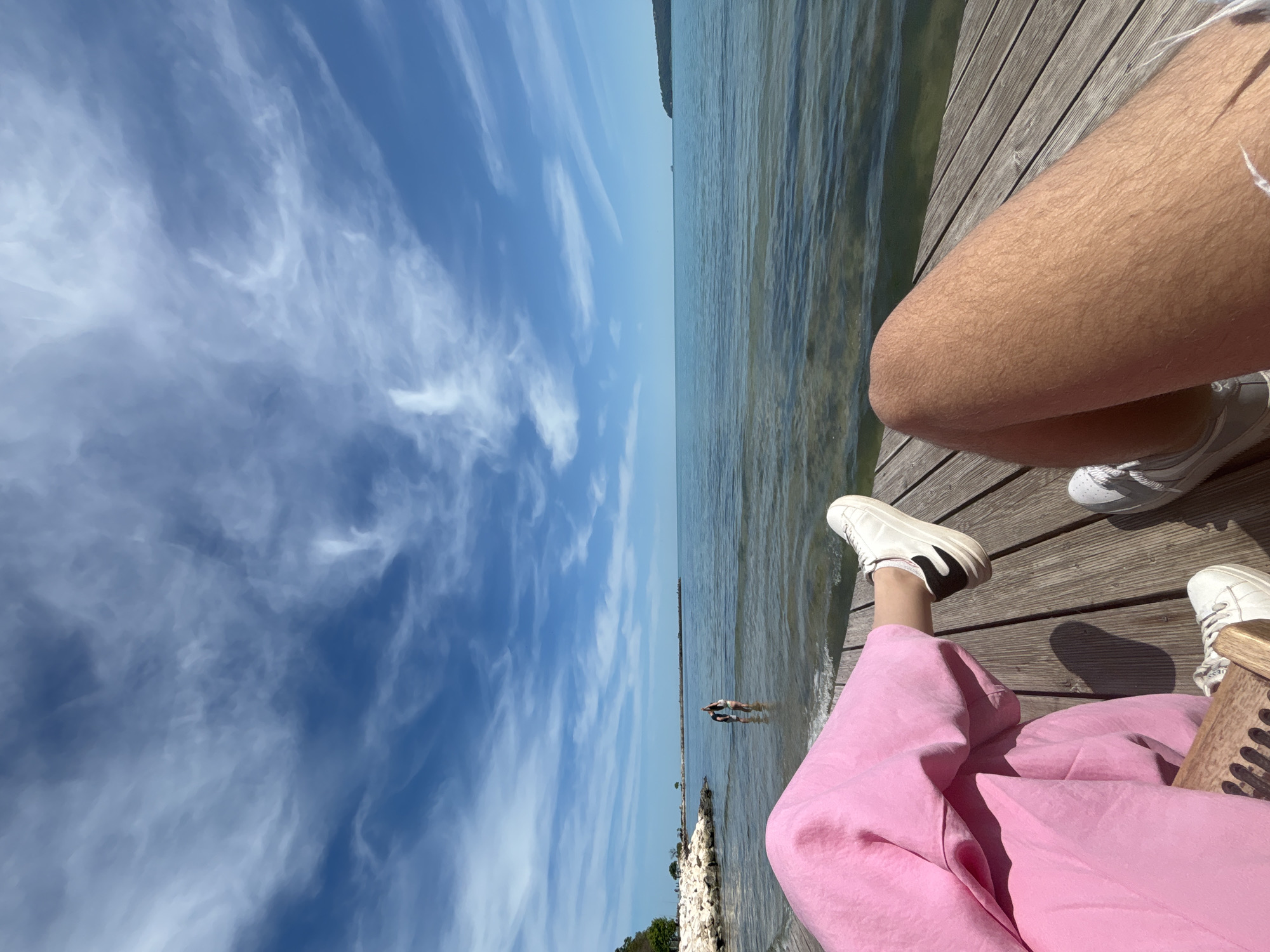 An eye-level shot, rotated ninety degrees, depicts two people on a wooden pier extending into the sea in Varna, Bulgaria. The sky above is light blue with wispy, white clouds. A body of water, presumably the Black Sea, is visible on the right side. In the water, there are two people standing far from the pier. On the pier, two pairs of legs and feet in white shoes are visible, belonging to two seated individuals. The person in the foreground wears pink trousers. A wooden object is situated next to them. The pier is constructed of light-colored wooden planks. In the distance, the coastline is discernible. The scene suggests a tranquil, sunny day at the seaside in Varna.