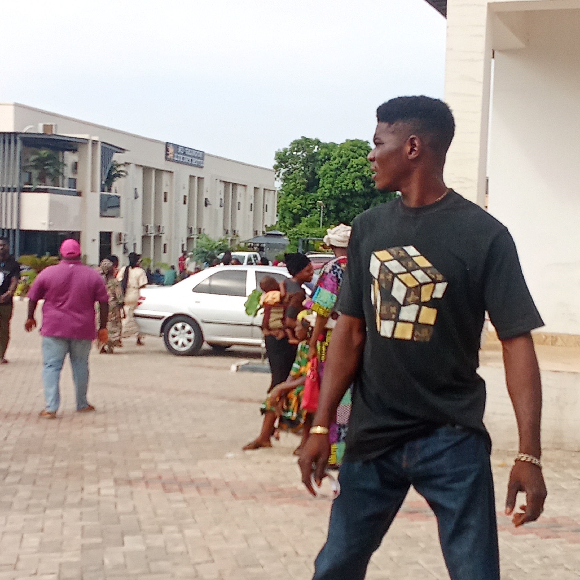 A street scene in Akwanga, Nigeria shows a young Black man wearing a black t-shirt with a Rubik's Cube-like graphic, dark denim jeans, and a gold chain bracelet. He is looking to his right. Several other people are in the background including a man in a pink hat, a woman carrying a child and others dressed in colorful clothing. A silver sedan is also in the background. A building is visible with a sign that reads "EL-RUFAI NURSERY AND PRIMARY SCHOOL." The scene takes place in daylight on a paved area.
