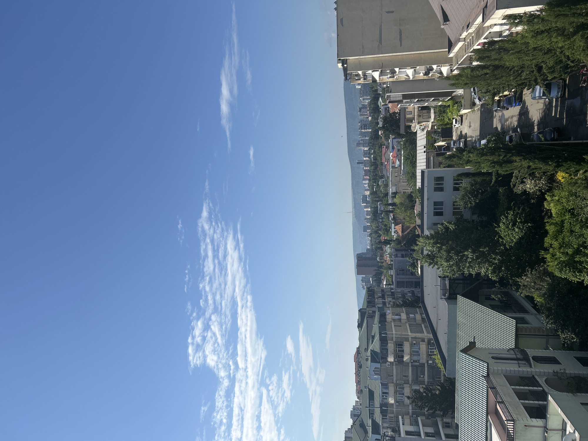 The image shows a view of a city with buildings and a street, under a bright blue sky with scattered white clouds. The buildings are of various heights and architectural styles, with some appearing to be residential apartment complexes and others smaller houses. Many buildings have green roofs. Tall, slender evergreen trees line the street, which is populated with several parked cars. In the distance, a larger cityscape is visible with taller buildings, and a range of hills or mountains forms the horizon. The overall impression is of a sunny day in an urban or semi-urban environment.