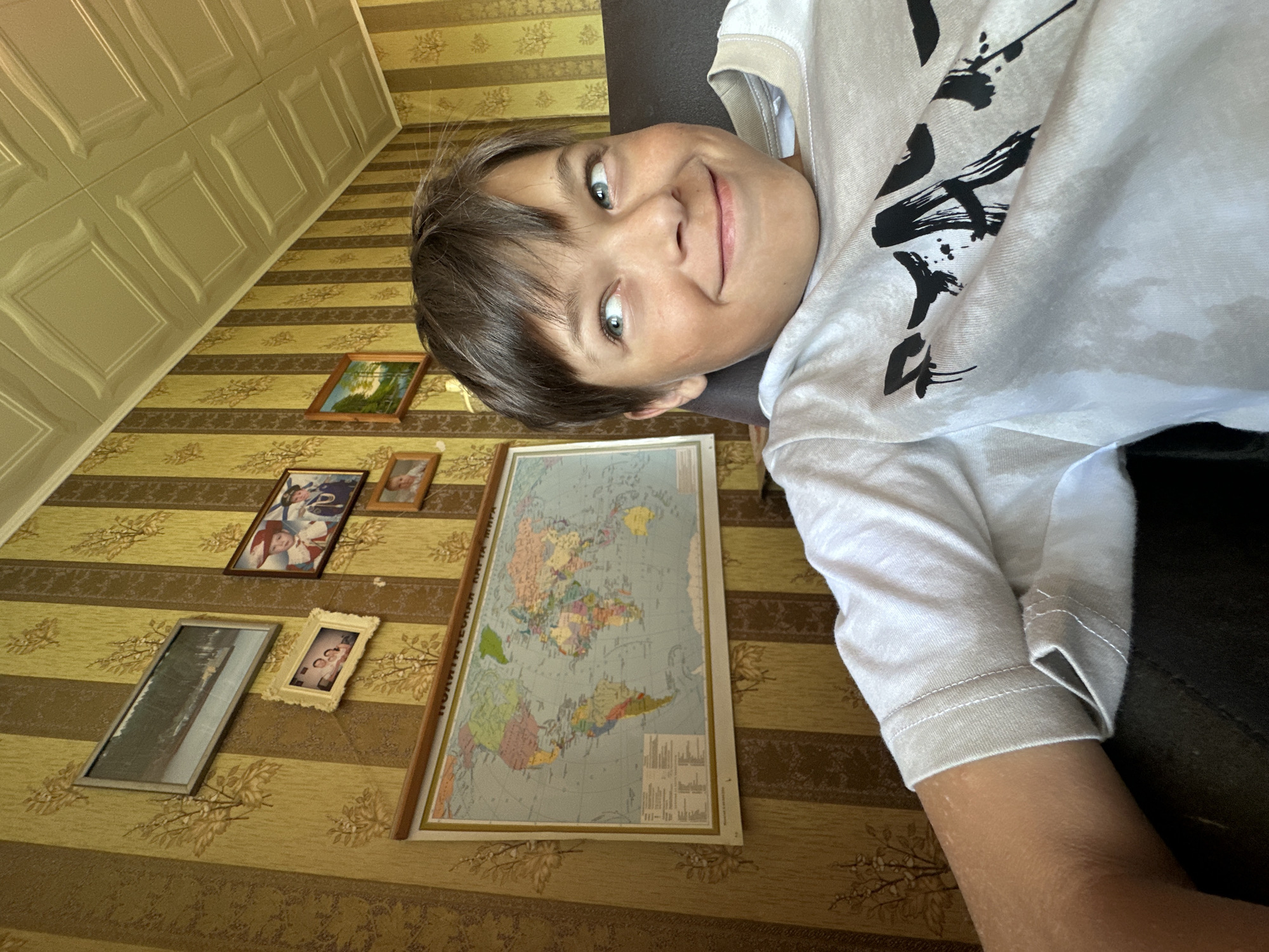 A young person with short brown hair and green eyes is lying down, facing the camera. They are wearing a white t-shirt with black Japanese-style lettering. Behind them, on a yellow and brown striped wallpapered wall, are several framed pictures and a world map. The location is Chaikovsky, Russia.