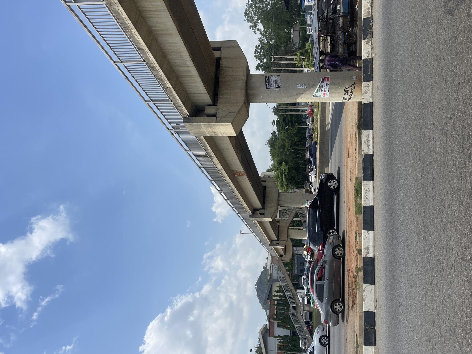 The image shows an elevated concrete structure with a metal railing, likely a pedestrian bridge or part of a transit system, spanning over a road in Gwarinpa, Nigeria. Several cars are parked along the side of the road, delineated by a black and white painted curb. A few people are visible near a pillar supporting the elevated structure. The sky is blue with scattered white clouds. Trees and buildings are visible in the background.