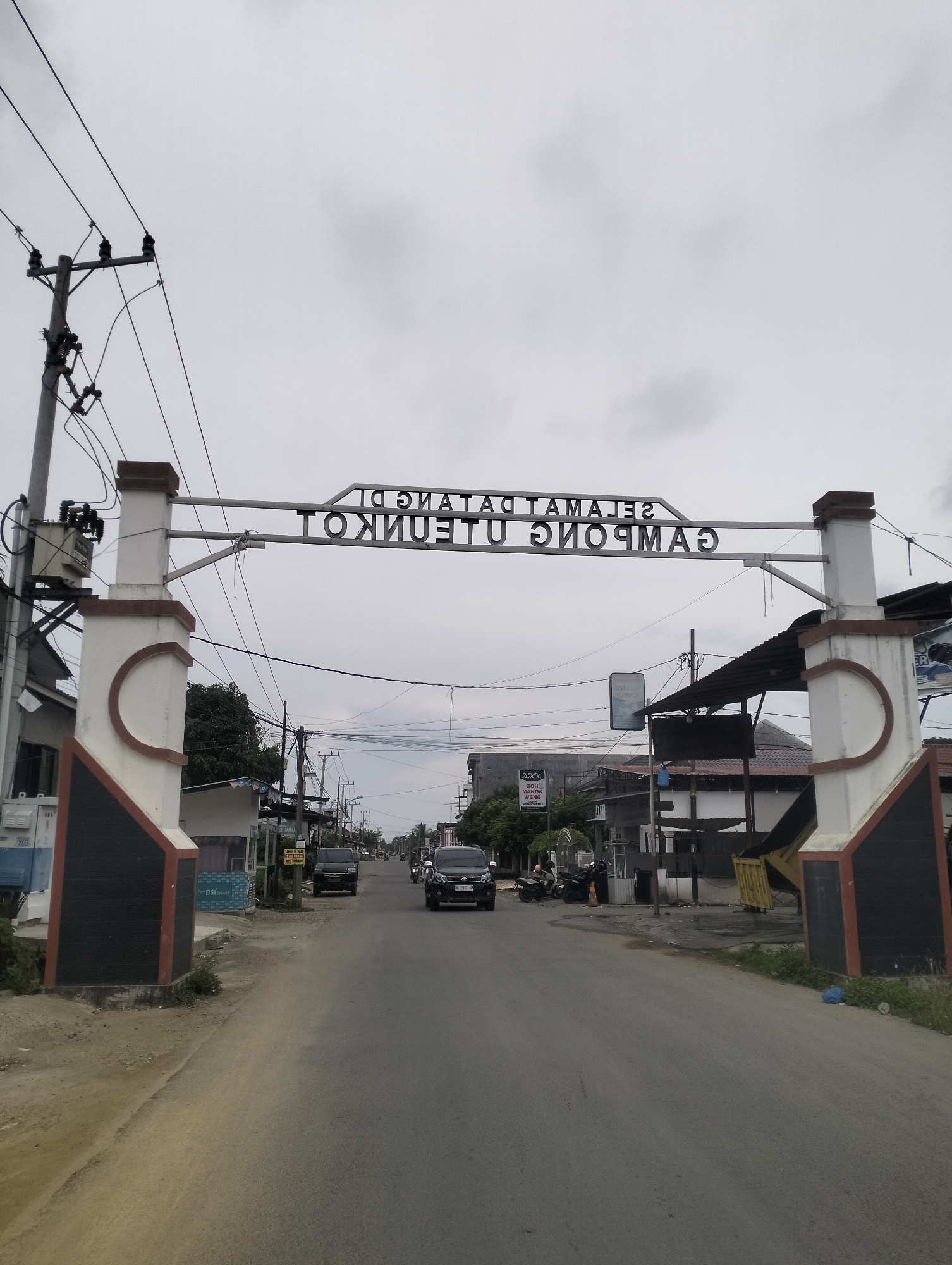 The image shows a street scene in Lhokseumawe, Indonesia, marked by an overhead welcome arch. The arch, inscribed with the words "SELAMAT DATANG DI GAMPONG UTEUNKOT," spans across the road. On either side of the entrance to the village are decorative pillars with brown and white accents. The road is paved and appears to be a main thoroughfare. Several vehicles are visible, including black SUVs, a motorcycle, and a parked van. Buildings line the sides of the street, some appearing to be shops or businesses, with awnings and signage. Utility poles with power lines are a prominent feature of the environment. The sky is overcast, suggesting a cloudy day. The overall scene depicts a typical street in a populated area of Lhokseumawe.
