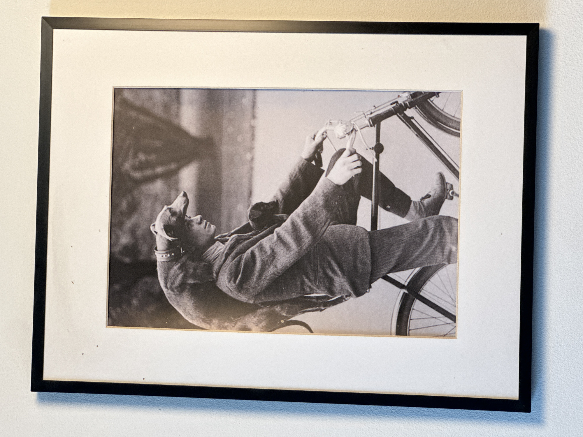 A black and white, framed photograph shows a person riding a bicycle with a dog draped over their shoulders. The person is wearing a tweed jacket and cap, and the dog is a sighthound breed. The background appears to be an outdoor setting with trees. The photograph is mounted on white matting and framed with a black border. The image is not located in Kuta Selatan, Indonesia.