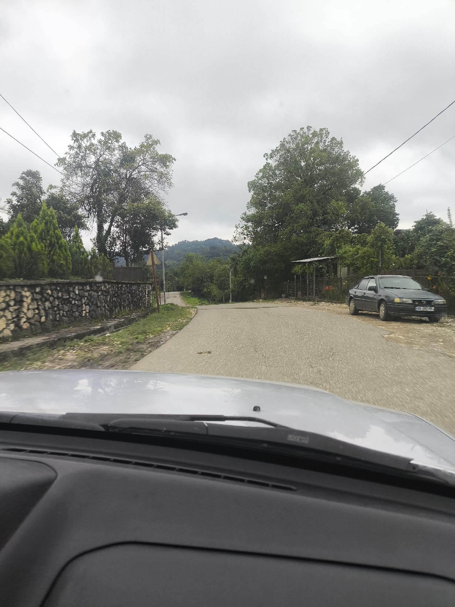 This image depicts a road in Skurdi, Georgia. The scene is viewed from inside a vehicle, looking out through the windshield and dashboard. A dark-colored sedan is parked on the right side of the road, facing away from the camera, with a license plate visible. On the left side of the road, there is a stone wall bordering a property with manicured coniferous trees. Further down the road, a paved surface transitions to a dirt or gravel area. Tall trees line both sides of the road, and a lamppost is visible. In the background, a hillside covered in vegetation suggests a rural or semi-rural setting. The sky is overcast and cloudy.