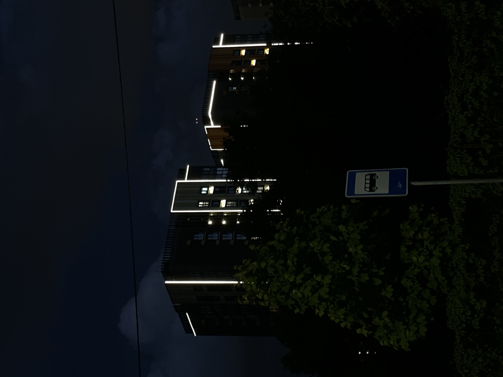 The image shows an urban scene at night in Minsk, Belarus. A tall residential building with illuminated horizontal and vertical light strips is visible. A bus stop sign stands on a pole, partially obscured by foliage. The sky is dark, with subtle cloud formations. Trees are interspersed throughout the scene.