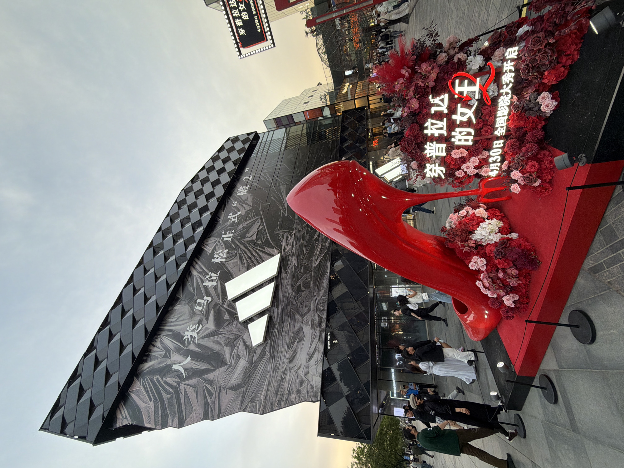 The image captures a vibrant street scene in Dongcheng District, China. A prominent Adidas store stands to the left, its facade featuring a combination of checkered black paneling and a gray textured surface adorned with the Adidas logo. A large, eye-catching red high heel sculpture is positioned in front of the store, decorated with an array of red and white flowers. A sign displays text in Chinese, promoting the film "The Queen Wears Prada". Several individuals are present, some standing near the flower-decorated display, possibly interacting with the exhibit, while others are seen walking in the vicinity. The overall environment suggests a busy urban setting with a focus on retail and promotion.