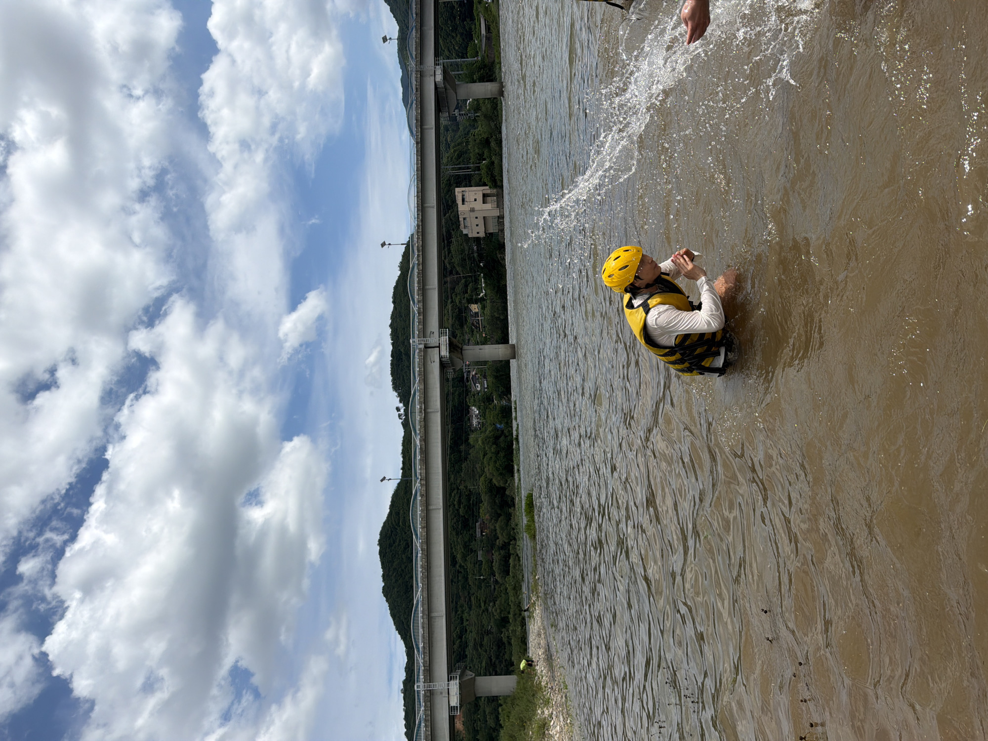 In the city of Seo-myeon, South Korea, a person wearing a yellow helmet and a yellow life vest is kneeling in a muddy river, splashing water. The person is wearing a white long-sleeved shirt under the life vest. The river is wide and appears to be shallow near the bank. In the background, a bridge spans across the river, supported by concrete pillars. Trees and hills covered with vegetation are visible beyond the bridge. The sky is partly cloudy with white clouds against a blue backdrop. The overall scene depicts an outdoor recreational activity on a warm day.