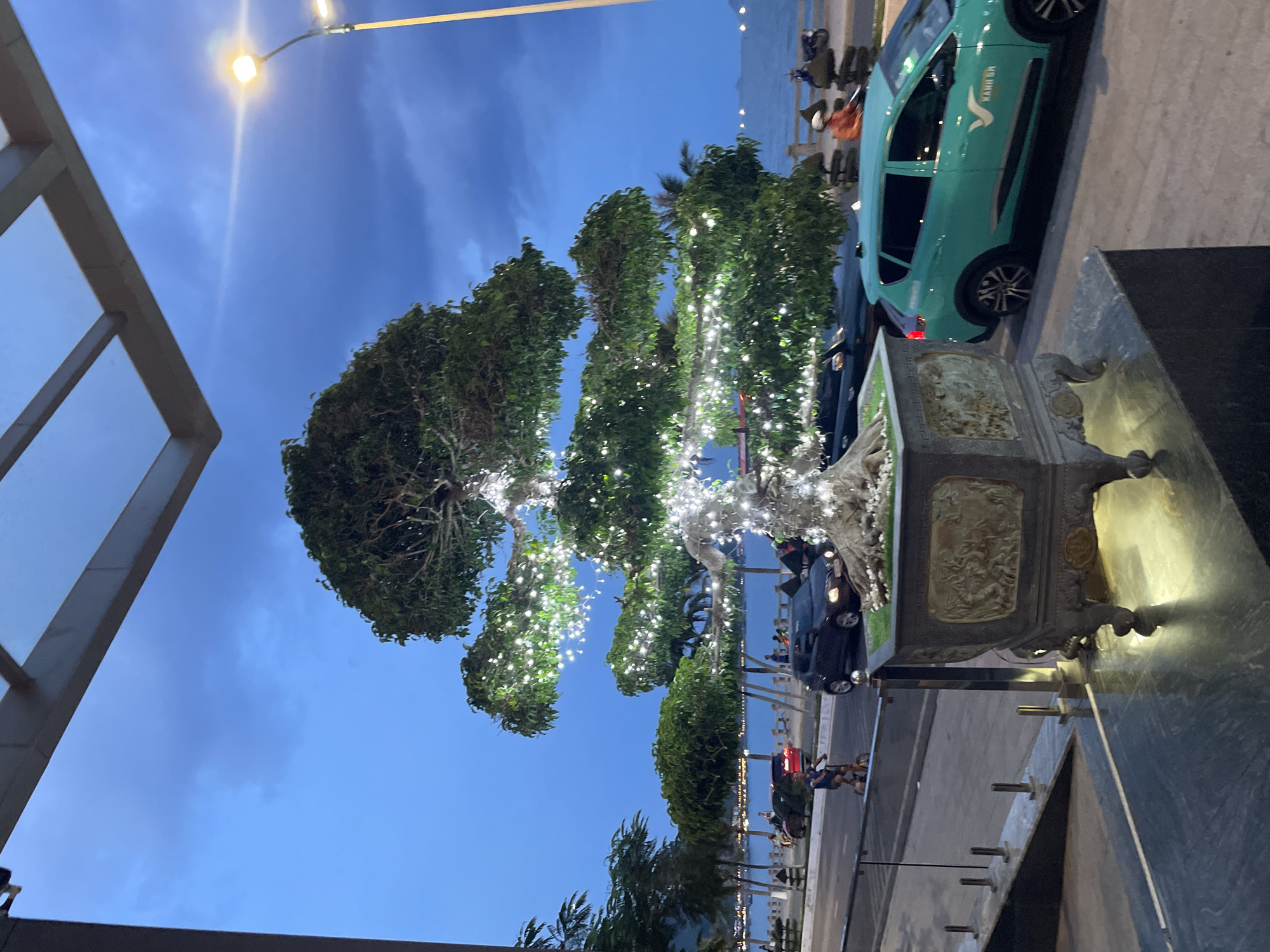 This is a vertical, medium-shot, eye-level photograph taken in Nha Trang, Vietnam, at dusk. The scene features a large, ornately potted bonsai tree adorned with numerous white fairy lights, illuminating its branches and trunk. The tree is positioned on a paved walkway next to a turquoise taxi.
In the background, a body of water, likely the sea, is visible, with a distant shoreline or islands. Several parked vehicles, including motorcycles and other cars, are present along the waterfront. A few people can be seen walking along the sidewalk and near the water. The sky is a deep blue, transitioning from daylight, and a street lamp is visible in the upper left corner, casting a warm glow.
The atmosphere is tranquil and festive, possibly indicating an evening event or a beautifully decorated public space. The ornate pot holding the bonsai tree is detailed with intricate carvings. Text is visible on the turquoise taxi: "KHANH HOA" and a stylized "V" logo. The overall impression is one of a peaceful, illuminated evening in a coastal Vietnamese city.