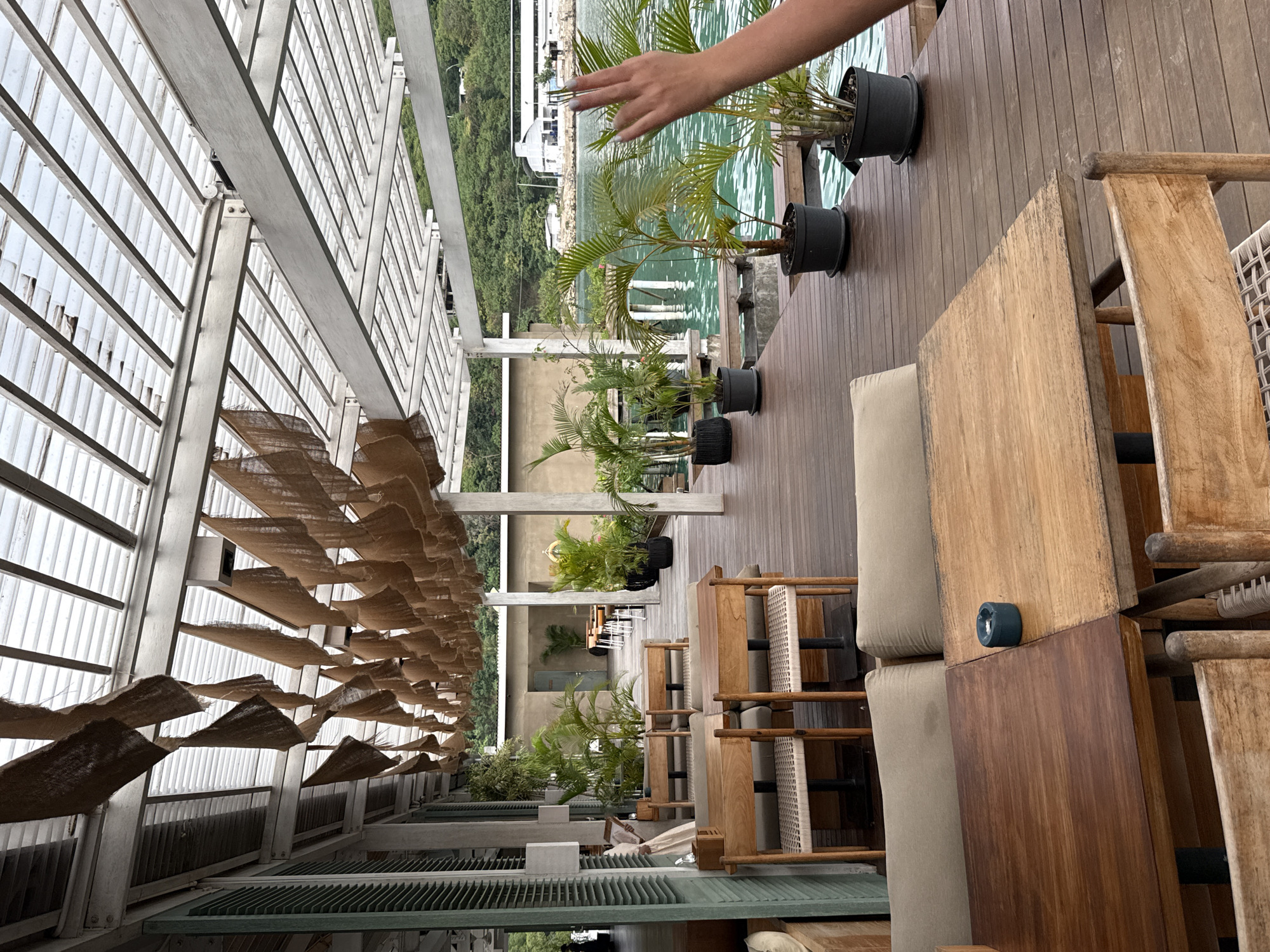The image shows an outdoor dining area in what appears to be a restaurant or lounge, likely in Labuan Bajo, Indonesia. The scene is captured from a slightly elevated, oblique angle. The dining area has wooden decking and is furnished with wooden tables, upholstered seating, and rattan chairs. Rows of potted palm trees line the edge of the deck, possibly overlooking a body of water; possibly the ocean, given the presence of green hills and building silhouettes in the background. Above the seating area is a structural canopy made of metal beams covered with panels with brown material. One arm reaches into the frame, suggesting that a person is either taking the photo or is engaged in an activity nearby. The scene appears to be set in a warm, tropical environment.