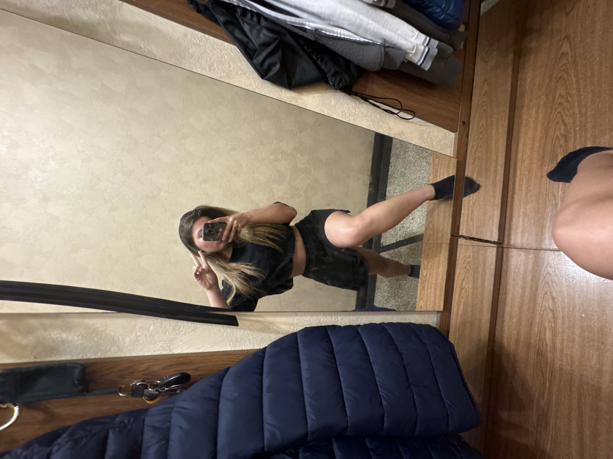 The image displays a mirror selfie taken by a person in an indoor setting, presented with a sideways orientation.
**Subjects:** The primary subject is a young woman, seen in reflection, taking the selfie. She has long, light-colored hair and is wearing a black short-sleeved crop top and dark shorts. Her face is mostly obscured by the smartphone she holds in her right hand. With her left hand, she forms a peace sign. Her right leg is extended and slightly raised, with a dark sock visible on her foot. A portion of another bare leg and a foot wearing a dark sock are visible outside the mirror's reflection in the bottom right corner, possibly belonging to the same person.
**Objects:** The central object is a large mirror reflecting the woman and parts of the room. A smartphone, likely an iPhone, with a clear case is visible in her hand. Above the mirror, a wooden shelf holds folded garments including black, grey, and white items. Below the mirror, a dark blue puffer jacket is draped over another wooden surface, alongside a dark green pouch and a set of keys attached to a ring. The room features light beige or off-white textured walls and wooden paneling or furniture elements.
**Scene and Setting:** The setting appears to be an interior room, possibly a changing room, hallway, or a bedroom with a large mirror and storage. The wooden elements and simple wall finish suggest a practical, possibly utilitarian space.
**Activity:** The primary activity is the act of taking a mirror selfie. The woman's pose, with an extended leg and a peace sign, indicates a casual and possibly playful moment.
**Notable Details:** The lighting suggests an indoor environment. The wooden textures are prominent throughout the scene. The image provides no specific time of day or weather cues. No visible text is legible in the image. The location context provided is Sofia, Bulgaria.
