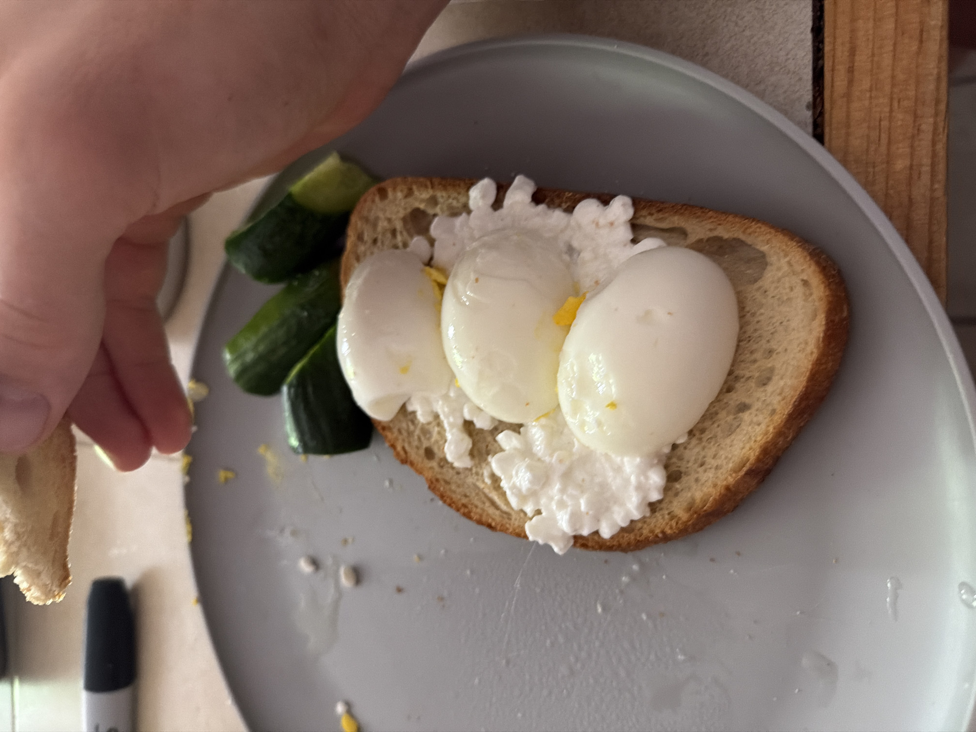 A hand reaches towards a gray plate containing a slice of bread topped with cottage cheese and three poached eggs. Cucumber slices are arranged on the side. The setting appears to be indoors, possibly a kitchen or dining area. A piece of bread is being held by the hand. A black marker is visible in the lower left corner of the plate. The scene is not identifiable as being in New York City.