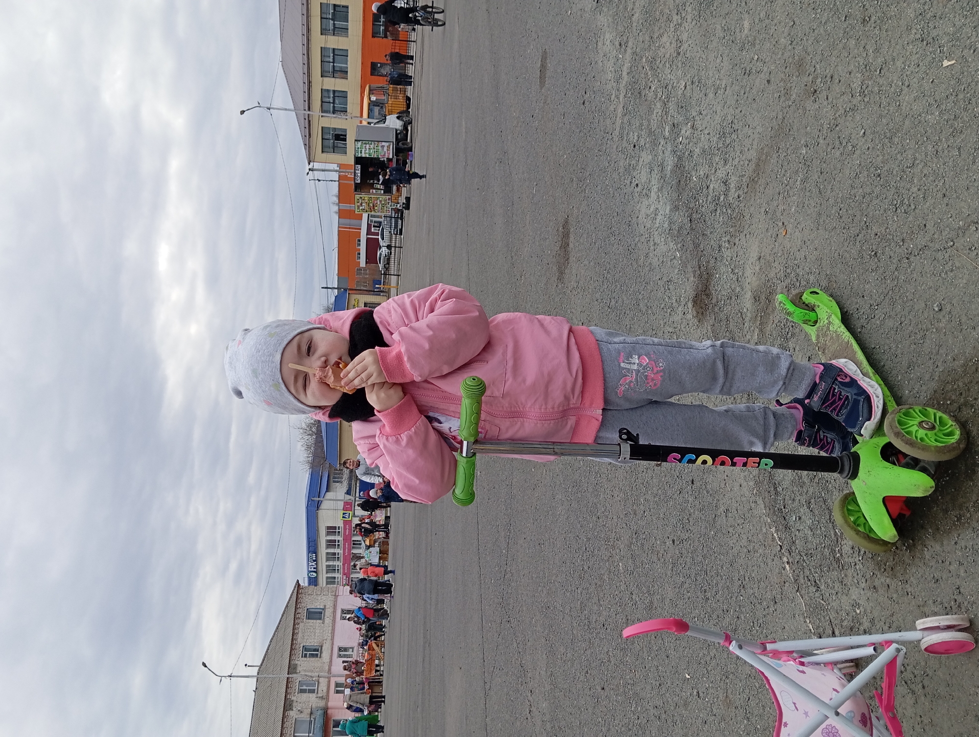 A young girl is eating a snack while standing on a green scooter in Yurgamysh, Russia. She is wearing a pink jacket, grey pants, a grey hat, and colorful sneakers. The scooter has black lettering that spells out "SCOOTER". A pink and white toy stroller is on the ground nearby. In the background are buildings, some with orange and tan facades, and a crowd of people in the distance. Overhead is a cloudy sky, and streetlights are visible along the street. The ground is a grey, asphalt-like surface.