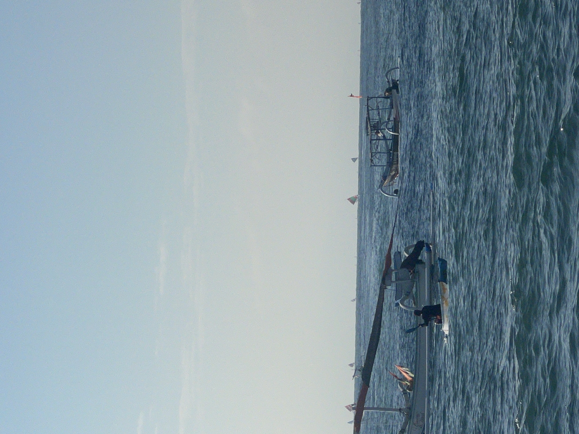 The image depicts a seascape with several boats on the water under a clear, pale blue sky. In the foreground, a traditional Indonesian outrigger boat, likely a jukung, with its outriggers extended, is positioned horizontally across the frame. A single person is seated in a kayak in front of this boat, paddling with a blue oar. Another jukung, with a thatched roof structure and a red flag, is visible further out on the water. In the distance, numerous smaller sailboats with triangular sails are scattered across the horizon. The water is choppy with visible ripples. The scene is likely Abang, Bali, Indonesia, a region known for its coastal activities and traditional fishing boats.