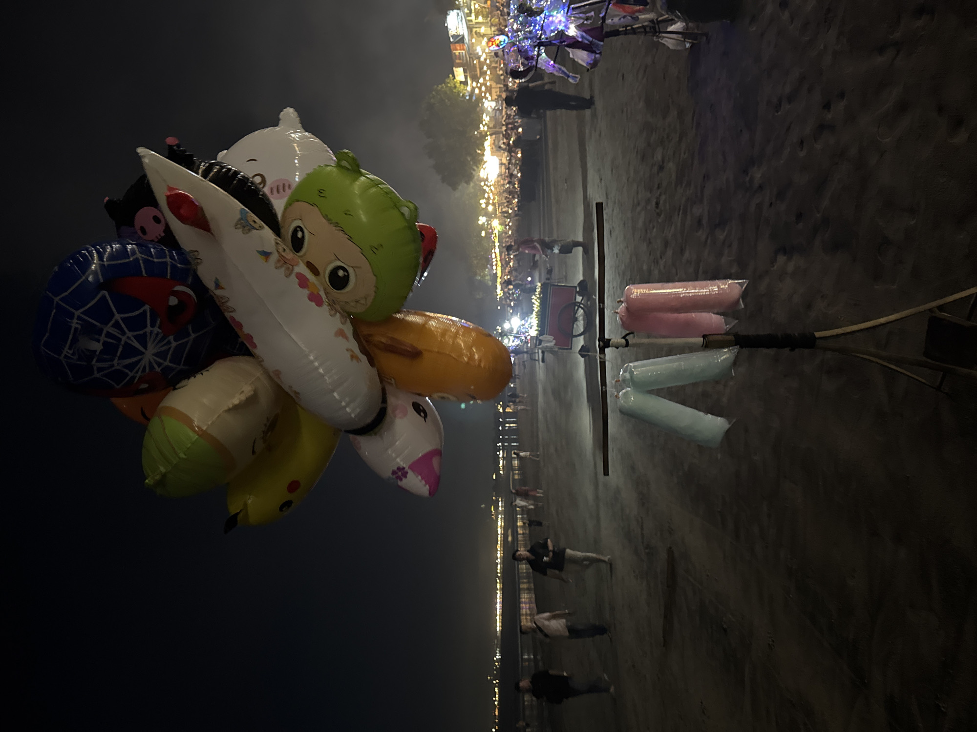 This nighttime image captures a vibrant outdoor scene in Kuta Selatan, Indonesia, likely on a sandy beach or promenade. The foreground is dominated by a large cluster of inflatable toys and balloons, varying in shape, color, and character. These include a blue Spiderman design, a green frog-like character, a white rounded animal, a yellow banana, and other cartoon figures, suggesting items for sale.
The setting features a wide, sandy ground with visible footprints, extending from the foreground into the mid-ground where people are strolling. In the mid-ground and background, a bustling area is brightly illuminated by numerous artificial lights, likely from stalls, string lights, and shops, creating a festive atmosphere. Silhouettes of people can be seen walking and gathering in this illuminated zone.
To the right side of the frame, a tall, dark metal stand holds several elongated, cylindrical inflatable objects, two in pink and two in light blue, which appear to be either decorative or additional items for sale by a vendor. A cart, possibly belonging to a vendor selling light-up toys or balloons, is also visible near the center-right. The scene conveys a lively, recreational environment, characteristic of a popular tourist spot or a night market by the sea. The time of day is clearly night, with a dark sky contrasting with the bright artificial lights. There is no visible text in the image.