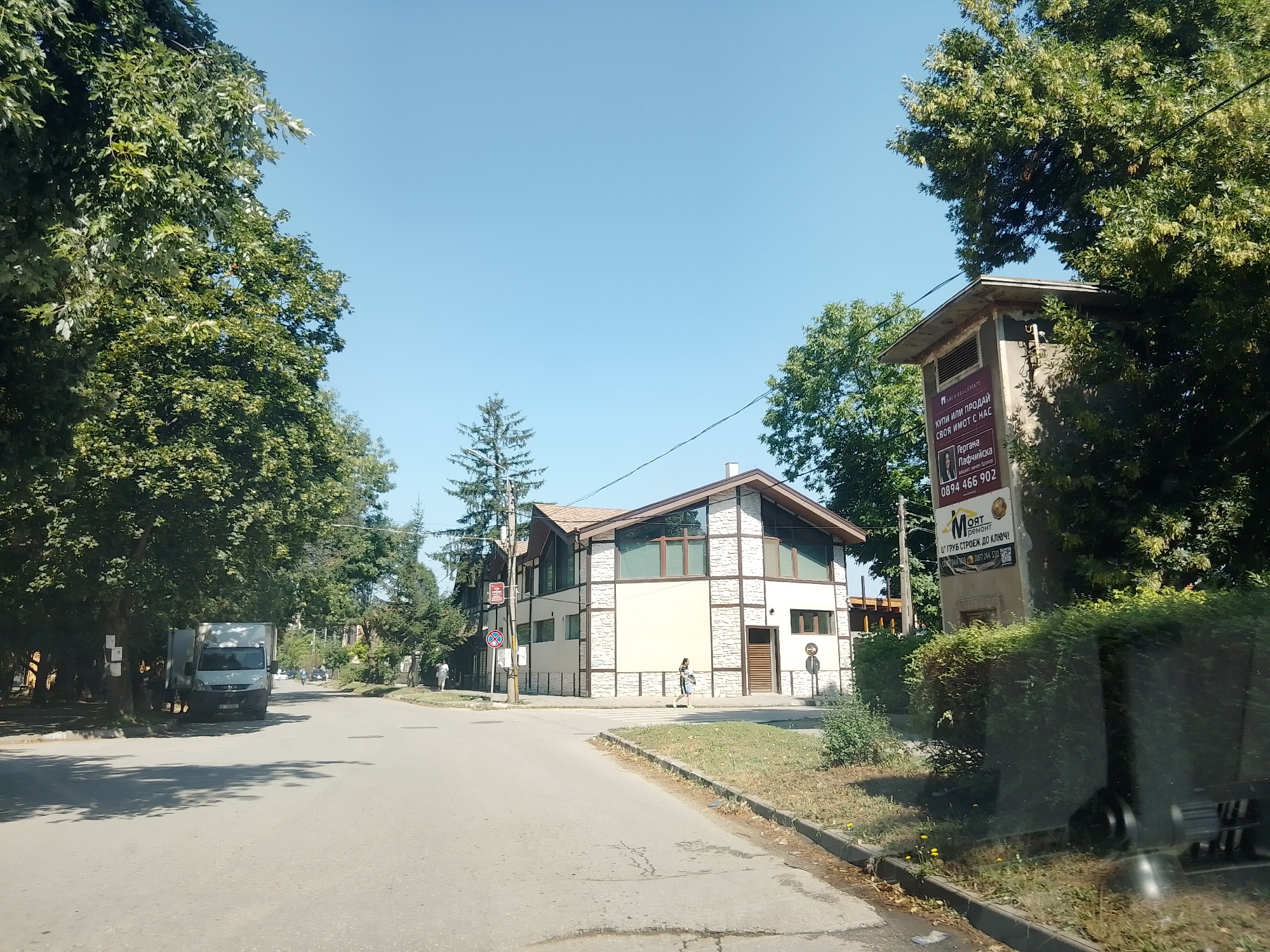 The image depicts a street scene in Ihtiman, Bulgaria, on a clear day. A light-colored van is parked on the left side of the road. Further down the street, a modern building with a stone and beige facade stands at an intersection. A pedestrian is walking across a crosswalk in front of this building. To the right of the building, a concrete structure with a sign advertising real estate services and renovation is visible. The street is lined with trees, and the sky is blue. The environment appears to be a suburban or small-town area.