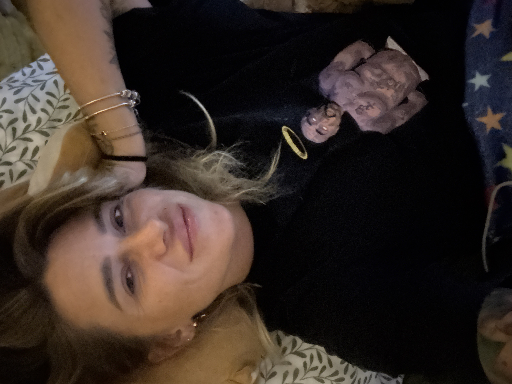 In this overhead, angled shot, a young woman lies on her side on a bed. She has blonde hair and is wearing a black top. She has several bracelets on her left wrist. In front of her, on the black fabric of her shirt, is a small, light purple statue of what appears to be a figure with a halo. The background is a duvet with a white and green leaf pattern. The lighting appears to be dim, suggesting it could be nighttime or indoors with low light. The woman's expression is relaxed and she seems to be looking directly at the camera.