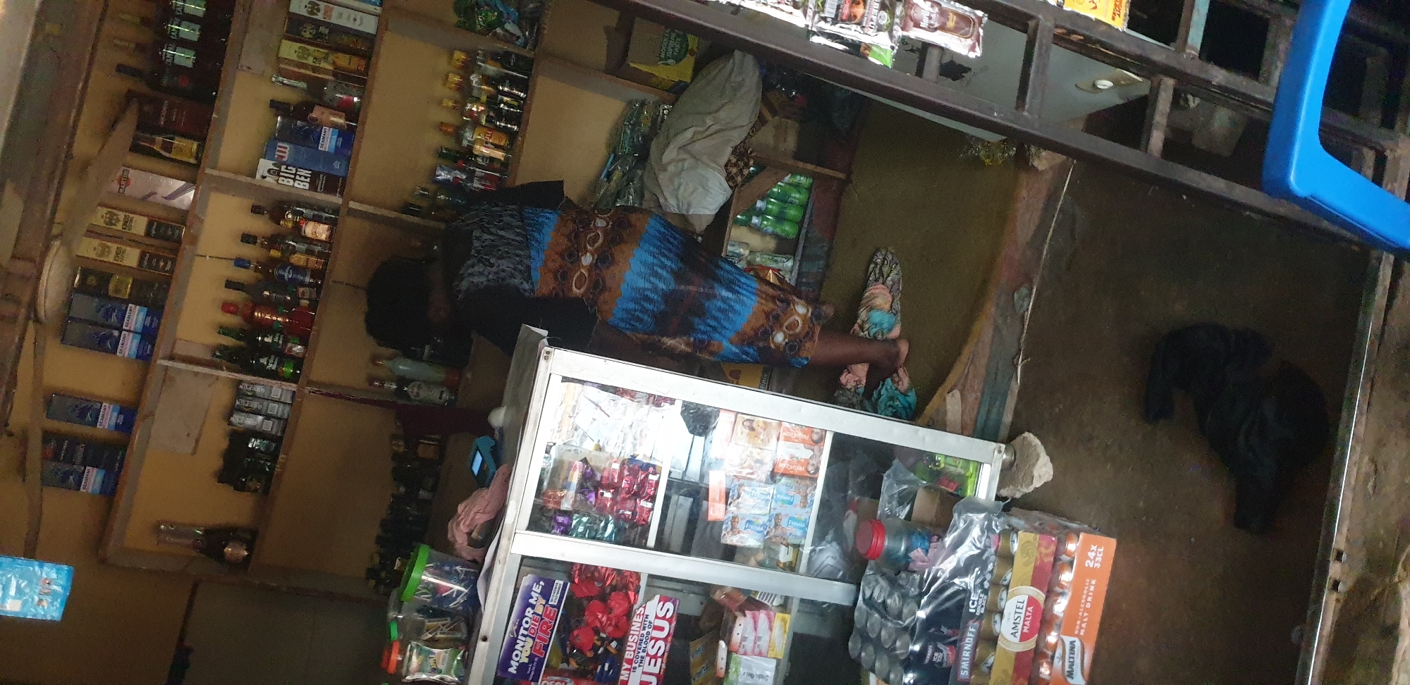 The image depicts the interior of a small shop, likely a convenience store or liquor store, in Sabon Tasha, Nigeria. The shop is densely stocked with beverages, including bottles of alcohol on shelves and stacks of canned drinks like Smirnoff, Amstel Malta, and Maltina in the foreground. Some items appear to be religious or motivational stickers on a display case. A woman is partially visible, wearing a patterned dress, and is interacting with a small electronic device, possibly a point-of-sale terminal. The environment appears somewhat cluttered, with goods stacked on shelves and on the floor, indicating a busy retail space. The lighting is dim, suggesting it could be evening or an indoor setting with limited natural light.