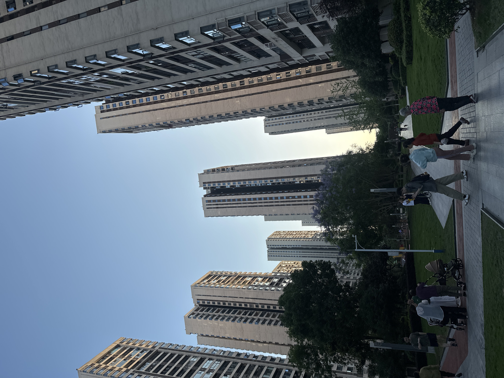 The image shows a view of the urban landscape of Chongqing High-tech Zone, China. Tall, modern apartment buildings with repetitive window patterns dominate the frame, angled upwards towards a clear, pale blue sky. A walkway, bordered by green lawns and trees, is visible in the foreground. Several groups of people are present: one group appears to be exercising, and another group is gathered near strollers.