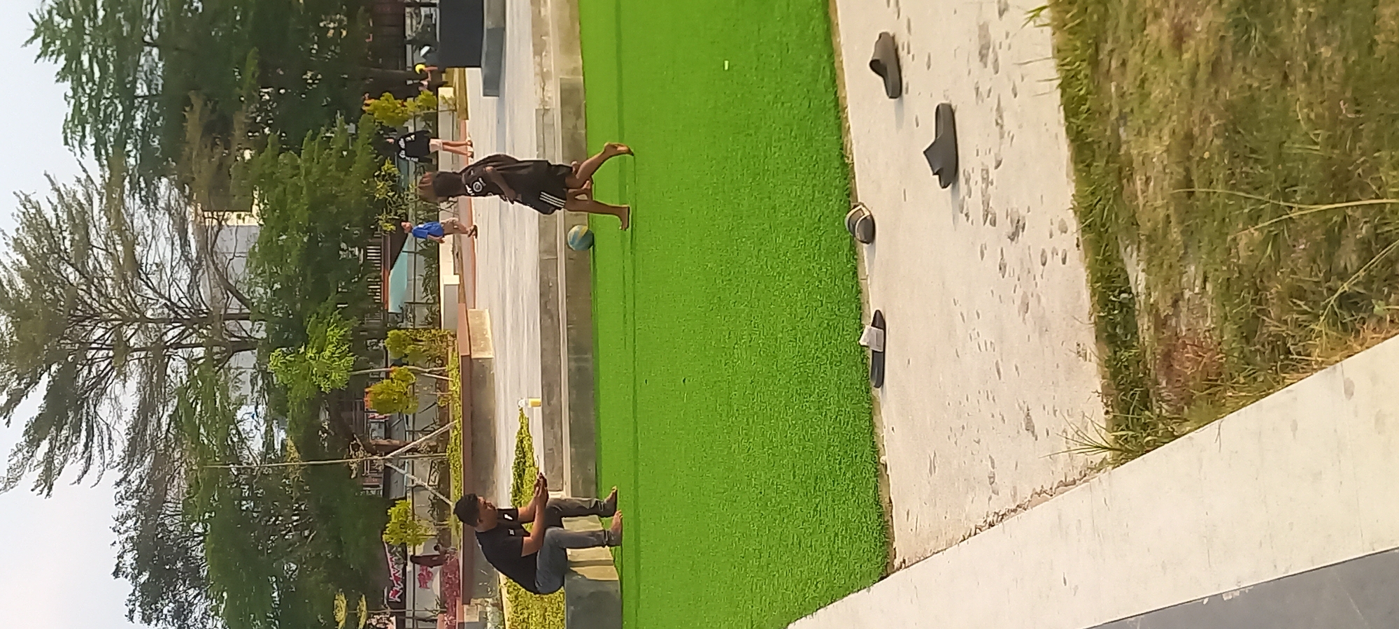 The image shows a group of people in an outdoor public space, possibly a park or recreational area, in Melayu, Indonesia. The foreground is dominated by a patch of artificial green turf, likely a sports field or play area. Several pairs of casual footwear, specifically flip-flops and sandals, are scattered on the concrete edge bordering the turf.
In the mid-ground, a man in a black t-shirt and grey pants is sitting on a concrete ledge, looking down at his phone. Beside him, a plastic cup containing a yellow liquid sits on the ledge. A few feet away, a young child in a black t-shirt and shorts is barefoot and appears to be playing with a soccer ball on the turf. In the background, there are trees, some buildings, and other people moving about. The overall lighting suggests it is daytime, likely late afternoon or early evening, given the soft light and shadows.