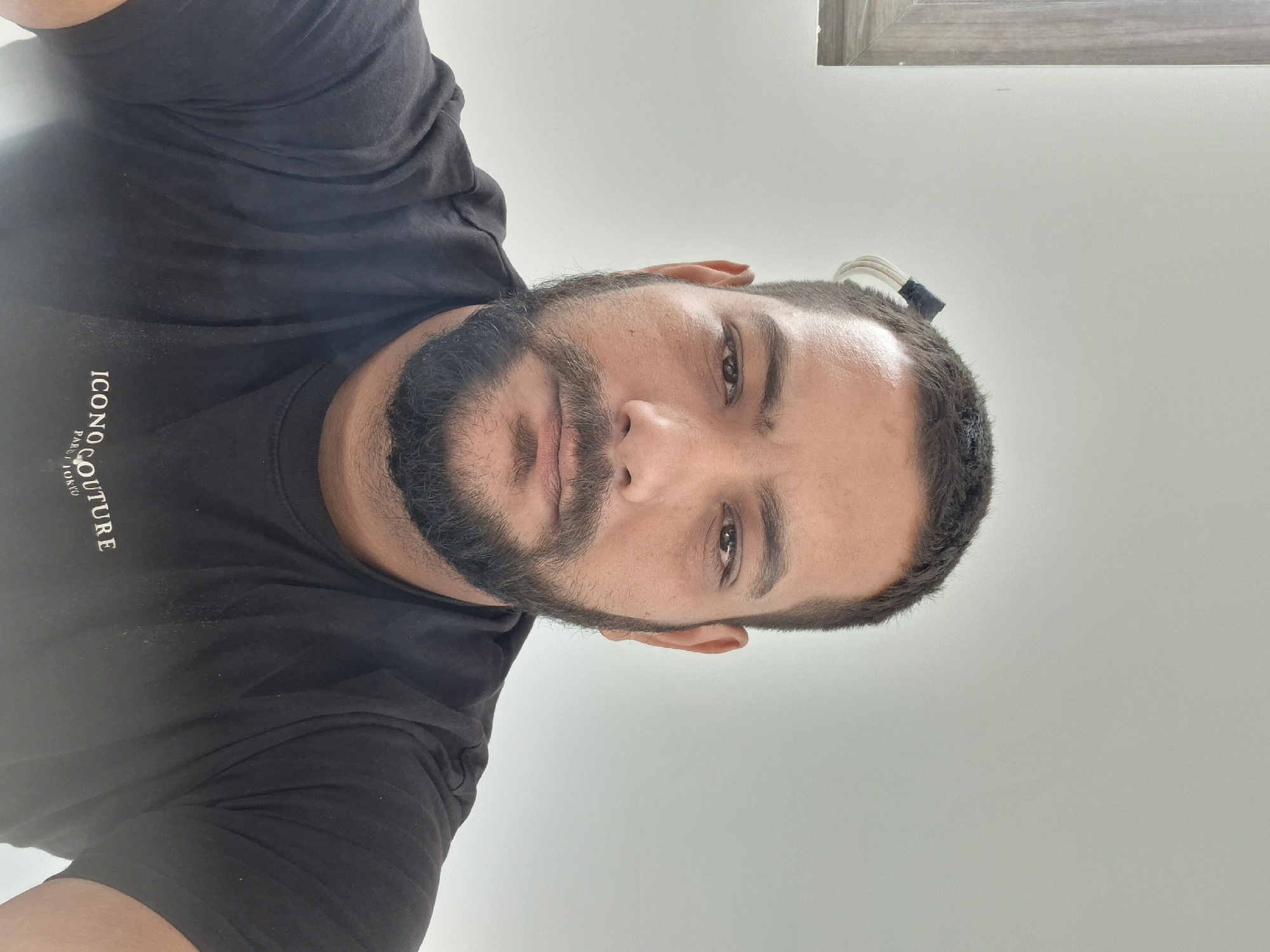 The image is a close-up, head-and-shoulders selfie of a man with dark hair and a full beard. He is wearing a black t-shirt with a white logo that reads "ICONOCULTURE PARIS TOKYO." The background is a plain, light-colored wall. There is a wooden frame on the left side of the image, suggesting the presence of a window or a piece of furniture. A small, black object with white wires is visible in the man's hair on the right side of his head. The man's expression is neutral. The image does not contain any specific visual cues to place it in Dolna Malina, Bulgaria; the description provided in the prompt is not reflected in the image.