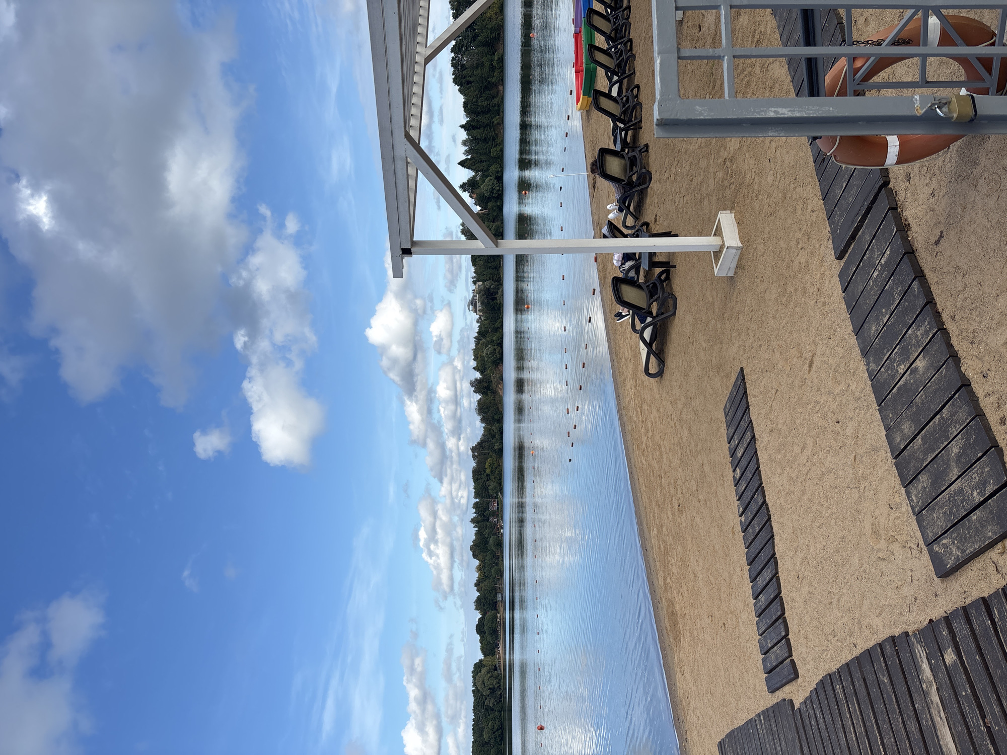 This is a scenic, eye-level, medium shot photograph taken at a lakeside beach in Рождествено, Russia. The foreground features a sandy beach with dark wooden boardwalks laid out in a crisscross pattern. Beyond the beach, a calm lake stretches out, reflecting the blue sky and scattered white clouds. A line of buoys marks a swimming area in the water. In the middle ground, several people are relaxing on lounge chairs under a white canopy structure. To the right, a metal gate with a locked padlock encloses a life preserver. The background is dominated by a dense line of green trees. The overall atmosphere is serene and tranquil.