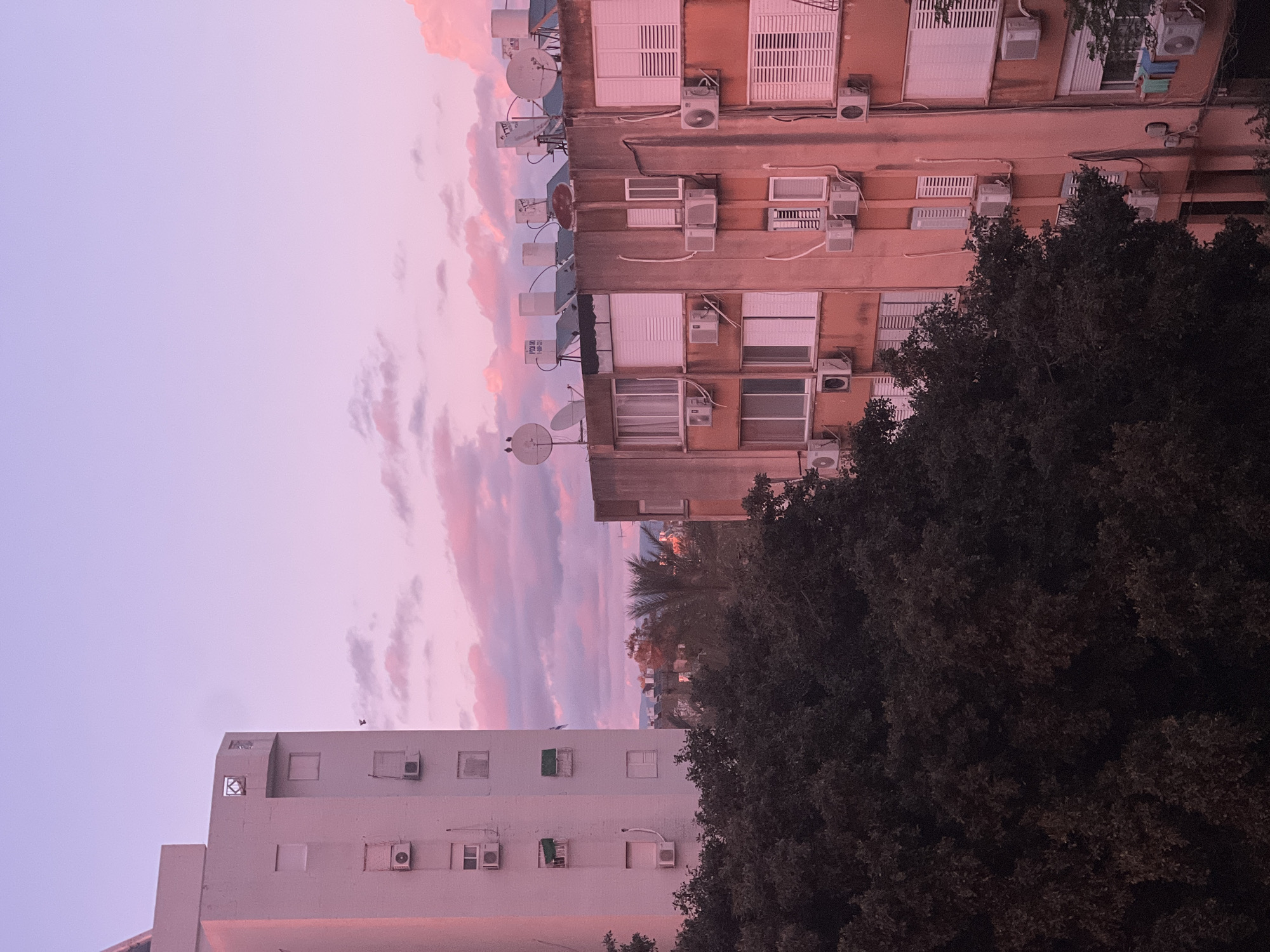 The image depicts a dusky cityscape in Kiryat Bialik, Israel, at dusk, oriented with the left side pointing towards the sky. The sky is filled with pink and purple clouds. Adjacent to the sky are the tops of two multi-story apartment buildings. The one on the left is white and has several air conditioning units on its facade. The building on the right is a faded salmon color, and features numerous air conditioning units and satellite dishes. A large, dark tree occupies the lower right quadrant. The perspective is a low-angle view.