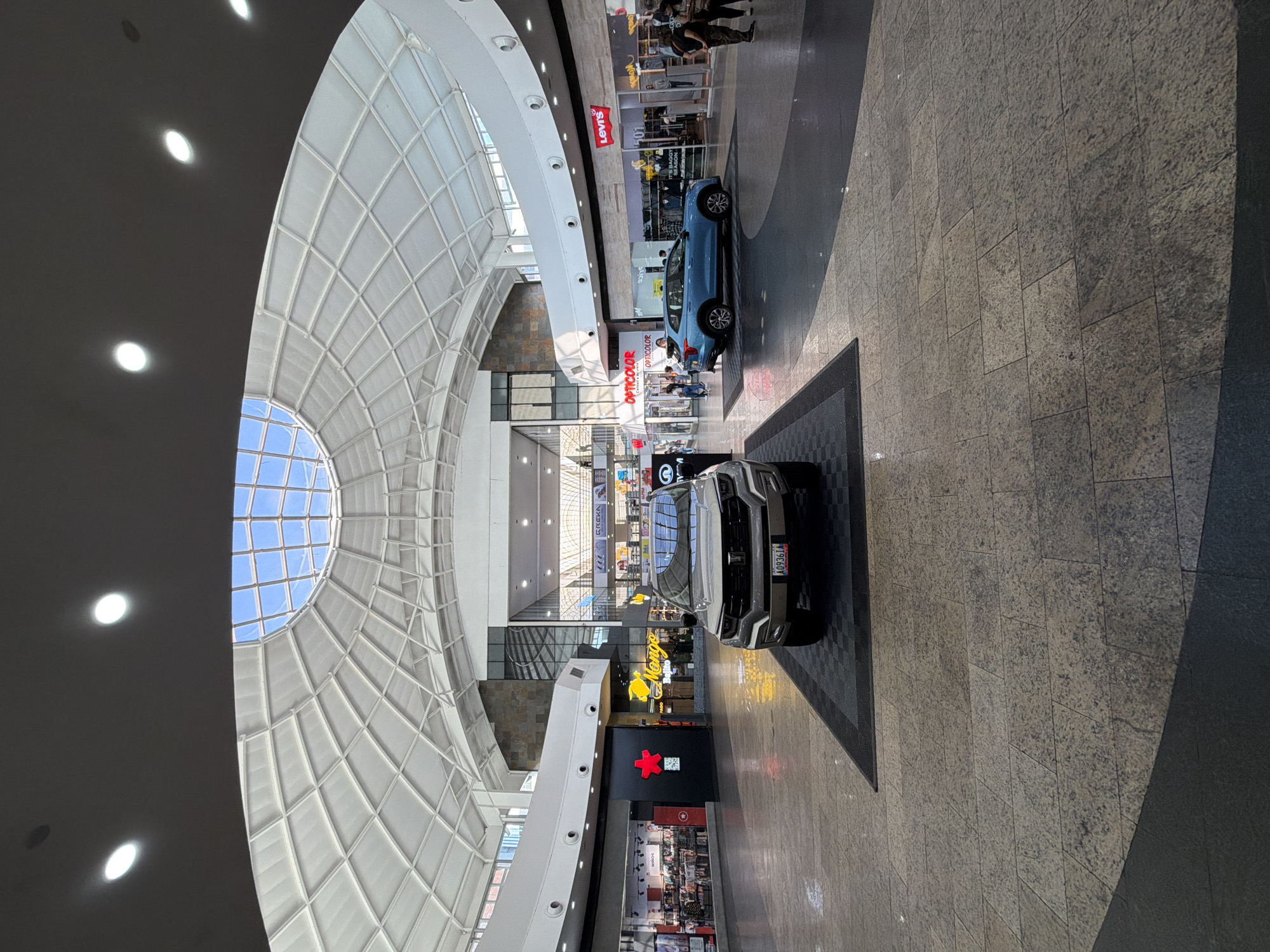 This image captures the interior of a modern, multi-story shopping mall, illuminated by both natural light and artificial fixtures. A prominent feature is the large, domed skylight with a grid pattern, allowing natural light from a clear blue sky to enter. On the left, the ceiling is dark grey with several circular recessed lights glowing brightly.
On the ground floor, which is paved with speckled light brown and grey tiles, two cars are on display. In the foreground, a silver or light grey SUV is parked on a dark checkered mat. Its license plate, "A09J61," is clearly visible. Further back, a light blue hatchback is also displayed, with a sign next to it bearing the "Opticolor" logo.
The mall features multiple storefronts on different levels. Visible store names include "Levi's" and "Opticolor" on an upper level, and "Yengo" and "Keka" on the ground floor. A distinct red cross-like emblem is also seen on a black storefront facade. A moderate number of people are present, seen walking, standing, and browsing near the stores and displayed vehicles. The scene depicts a typical daytime atmosphere in an active commercial setting. This shopping mall is located in Ciudad Guayana, Venezuela.
