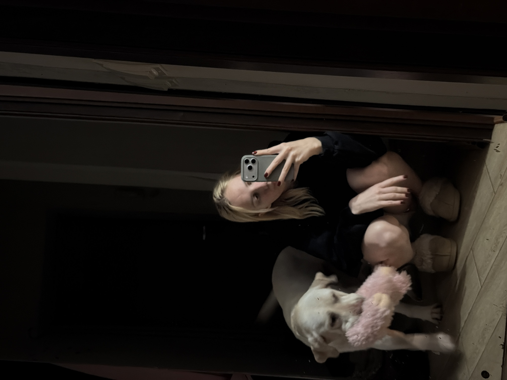 In Cheltuitori, Moldova, a dimly lit indoor scene captures a blonde female taking a mirror selfie with a smartphone, crouching on a tiled floor. She wears a dark long-sleeved top and light-colored fluffy slippers, with dark red nail polish visible on her right hand holding the phone. To her left, a light-colored Labrador-type dog is also low to the ground, holding a pink, fluffy toy in its mouth. The background is dark, with a reflective wooden door frame visible on the right.