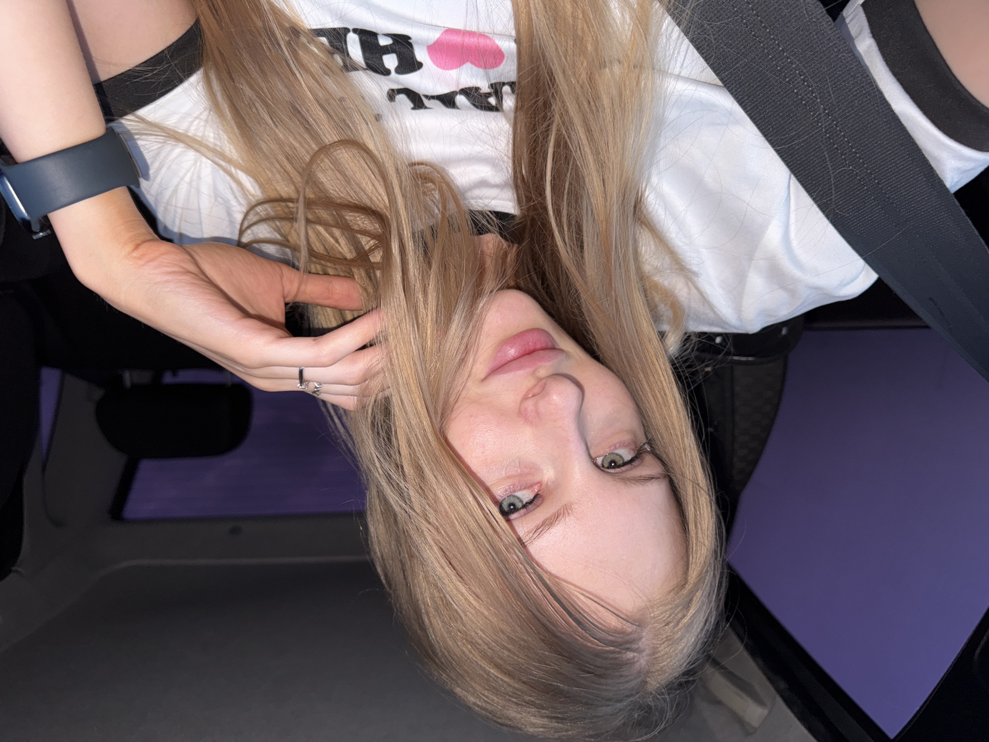 A young woman with long blonde hair and green eyes is taking a selfie inside a car. She is wearing a white t-shirt with black trim and a graphic that includes a pink heart and lettering. She has a seatbelt across her chest and is wearing rings on her fingers and a dark smartwatch on her wrist. The background shows the interior of a car with a window revealing a dark purple sky. The image was taken in the city of Perm, Russia.