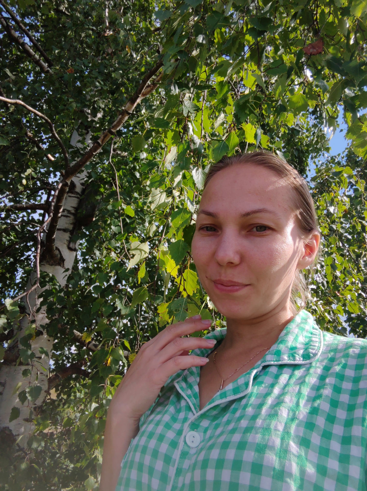 The image is a selfie taken outdoors, with a woman posing in front of a tree. She is wearing a green and white checkered shirt. The woman has light brown hair pulled back and is looking at the camera with a slight smile. She is touching her face with her left hand. The background consists of lush green leaves and branches of a birch tree, with a hint of blue sky visible. The setting suggests a natural environment, possibly a park or a backyard in the city of Chukhloma, Russia.