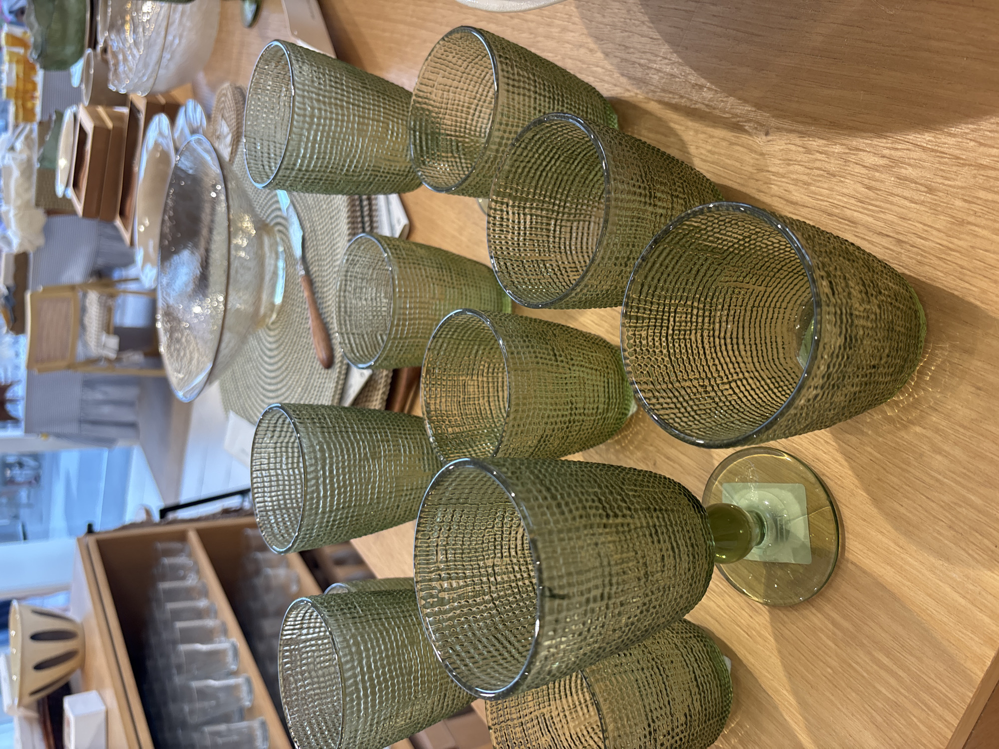The image shows a collection of green, textured glassware on a light brown wooden surface. There are several tumblers and one stemmed glass. In the background, a wooden shelf unit displays more glassware and a bowl. Further back, a chair and part of a table are visible. The scene appears to be a retail or display setting, possibly a homeware store or a kitchen. There are no people or animals present. The lighting suggests an indoor environment, possibly with natural light from a window. The image does not contain any information that would indicate its location in Orihuela, Spain.