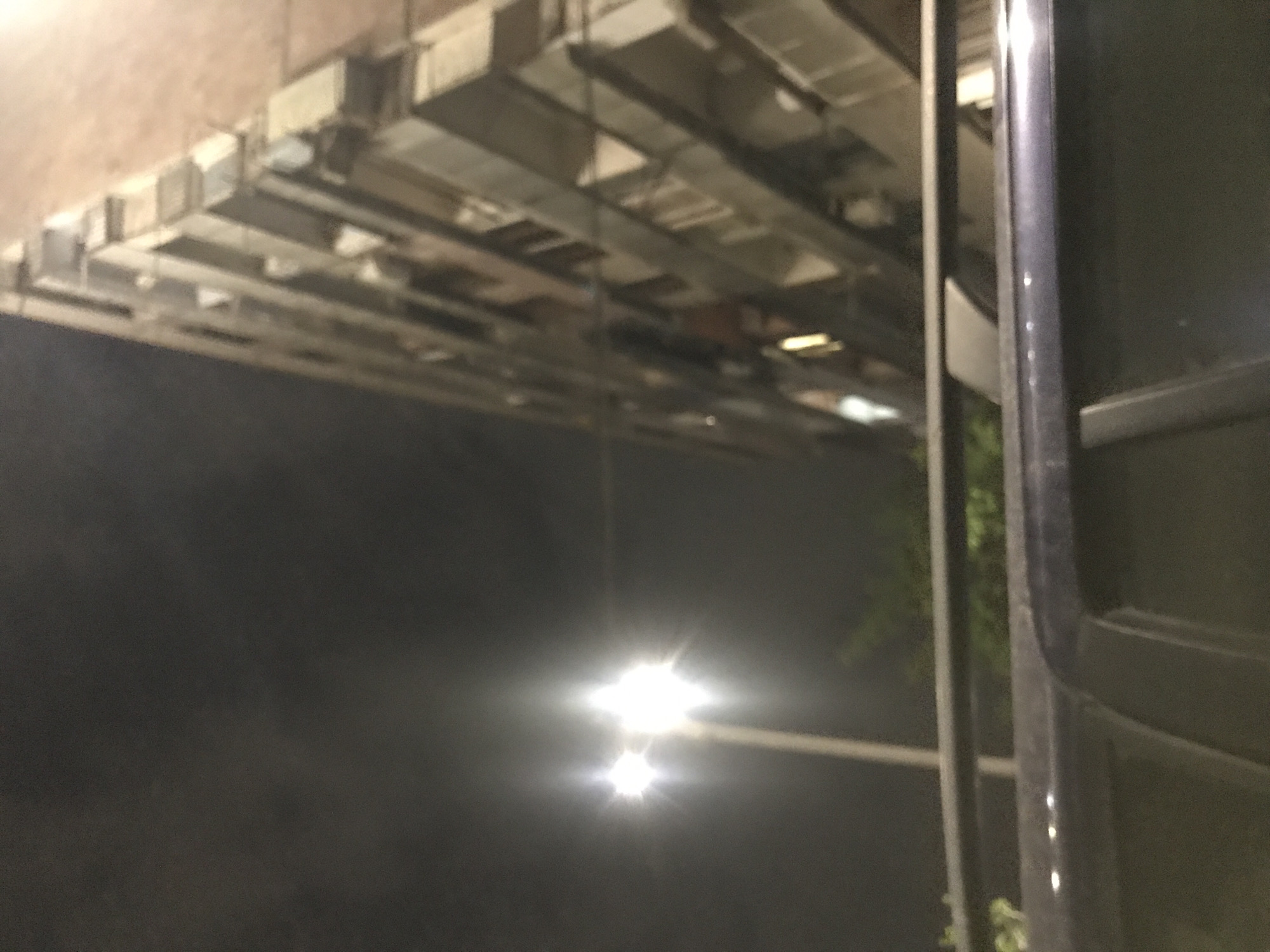 The image appears to be taken at night in an urban environment. Above is a ceiling with a grid-like structure. In the foreground is a dark object that may be part of a vehicle or building, possibly casting a shadow. A streetlight is visible in the distance, emitting a bright light. Due to the limited visual information available in the image, specifying the exact location within Yerevan, Armenia is not possible. The absence of people in the image suggests it could be an isolated or quiet area.