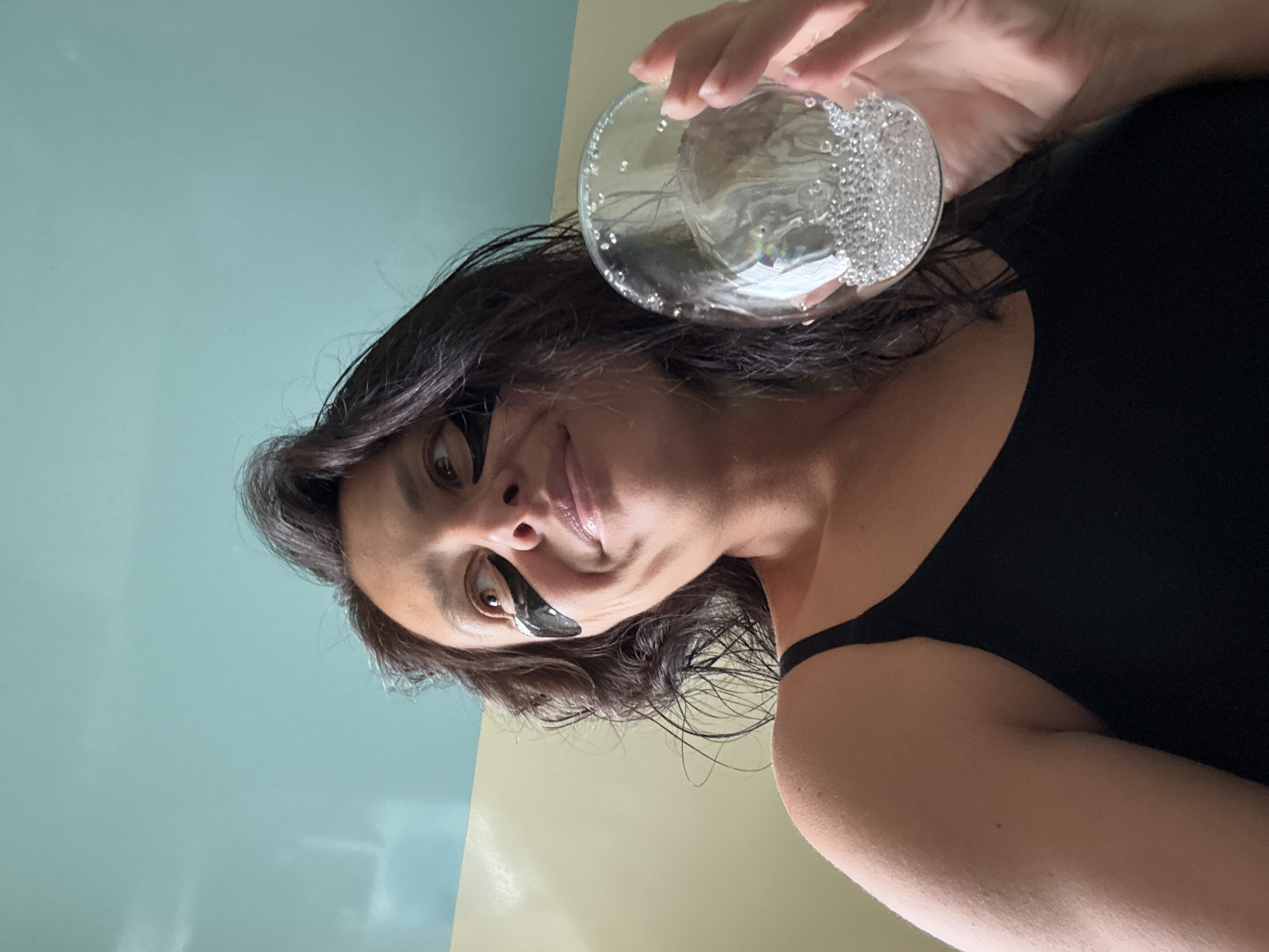 The image displays a close-up, low-angle perspective of a woman indoors, taken from a rotated position (90 degrees clockwise). The woman, with dark, wavy hair, is seen looking towards the camera with a relaxed expression and a gentle smile. She has a black, crescent-shaped under-eye patch or mask under her left eye, indicating a moment of self-care.
Her right hand extends into the upper part of the frame, holding a clear glass. The glass contains a liquid, likely water, along with numerous small, sparkling, spherical elements that appear to be decorative beads or bubbles. She is wearing a simple black top, partially visible on her shoulder and upper chest. The background consists of a plain wall, distinctly divided into two colors: a light blue/teal in the upper left portion and a light yellow/beige in the lower right.
The setting appears to be a private indoor space, suggesting a personal and calm moment in Chaikovsky, Russia. There is no visible text in the image.