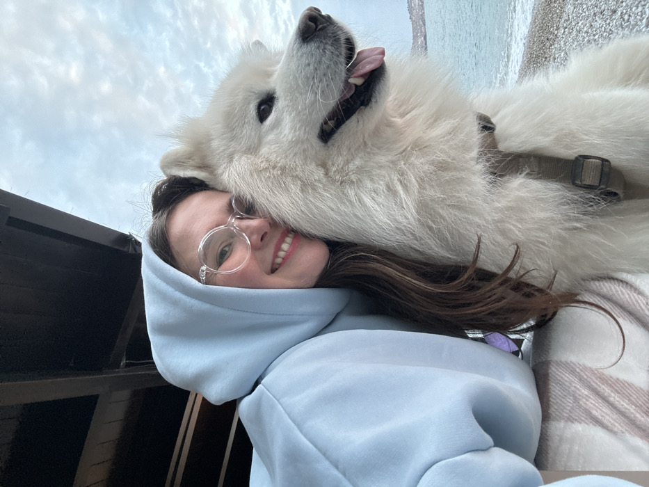 A female subject and a large white dog are depicted in a close-up, waist-up composition. The female, positioned on the left, wears a light blue hooded sweatshirt with the hood up, clear round-rimmed glasses, and has brown hair. She is smiling, revealing her teeth, and her head is pressed against the dog's head. The dog, a large, fluffy white breed consistent with a Samoyed, is on the right. It wears a beige webbing harness, has its mouth open, and its tongue partially extended. The dog's gaze is directed slightly off-frame to the right. A light-colored, striped fabric is partially visible beneath the female's left arm. The upper left background reveals dark wooden structural beams. The upper right background consists of an overcast sky. The mid-right and lower right backgrounds display a body of water, a pebbled shoreline, and distant landmasses on the horizon. The scene is situated in the city of Šušanj, Montenegro.