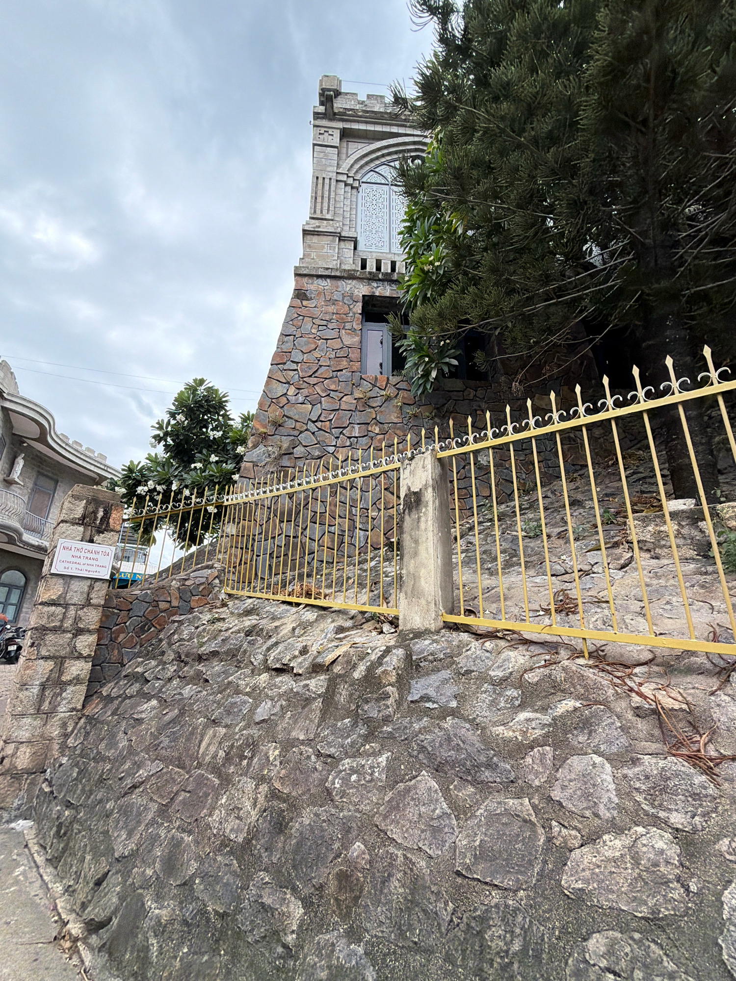 The image shows a view looking up at the Nha Trang Cathedral, a French Gothic-style building located in Nha Trang, Vietnam. The cathedral features a tall, stone tower with arched windows and intricate detailing. In front of the cathedral, a yellow metal fence runs along a steep, rocky slope. A sign on a stone pillar reads "NHÀ THỜ CHÁNH TÒA NHATRANG CATHEDRAL OF NHA TRANG 381, Thai Nguyễn". A large tree with dark green foliage partially obscures the view of the cathedral. The sky is overcast with light gray clouds. The overall scene suggests an overcast day in a somewhat humid environment. There are no people or animals visible in the image.