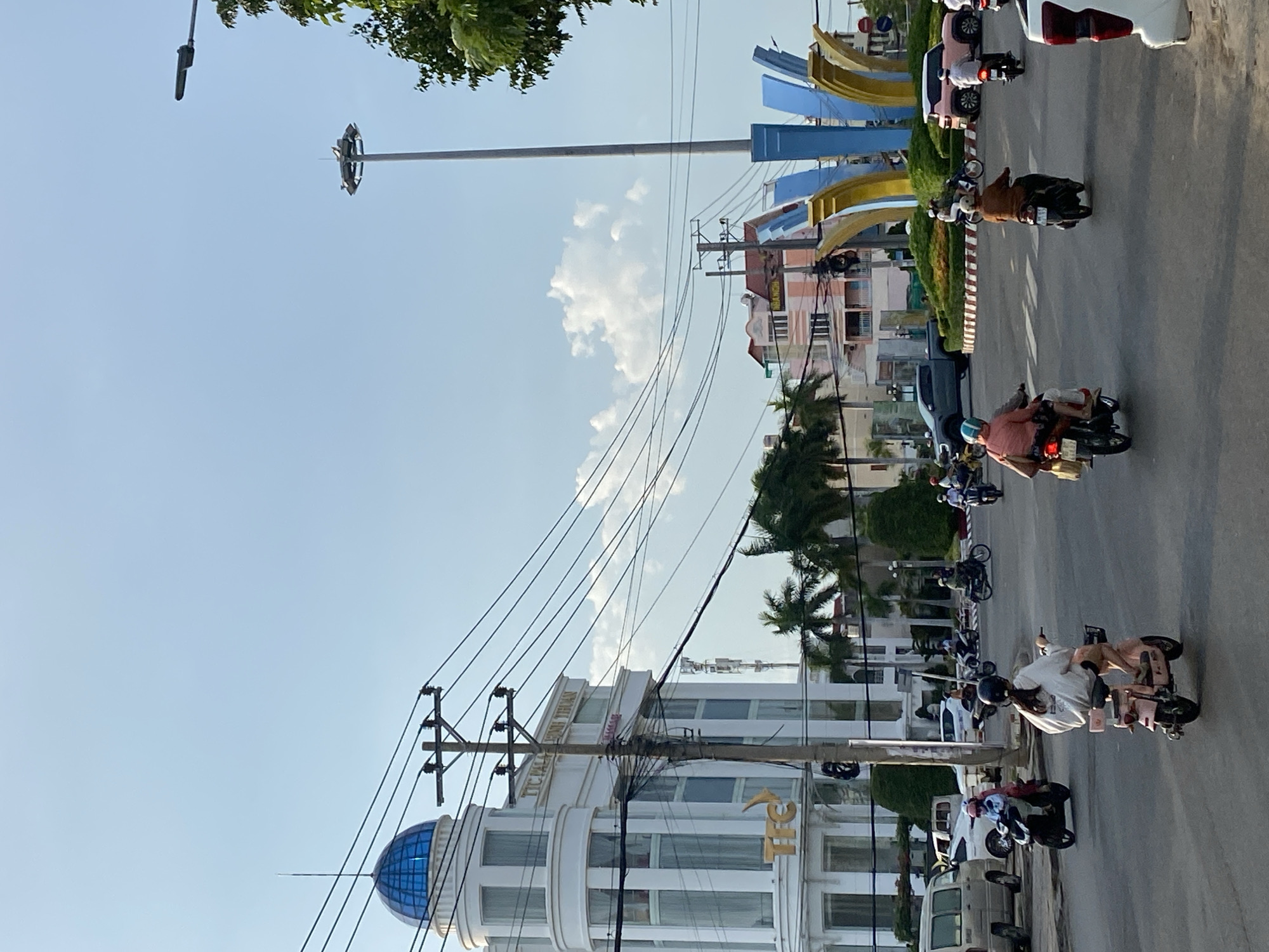 The image shows a street scene in Phan Thiết, Vietnam. The sky is clear with some clouds. A street with multiple motorbikes carrying people is visible. On the left side of the street is a large white building with a blue dome. Power lines and utility poles are strung across the sky. There are other buildings and trees in the background. There is a tall pole in the middle of the image. A monument consisting of blue and yellow curved structures is visible.