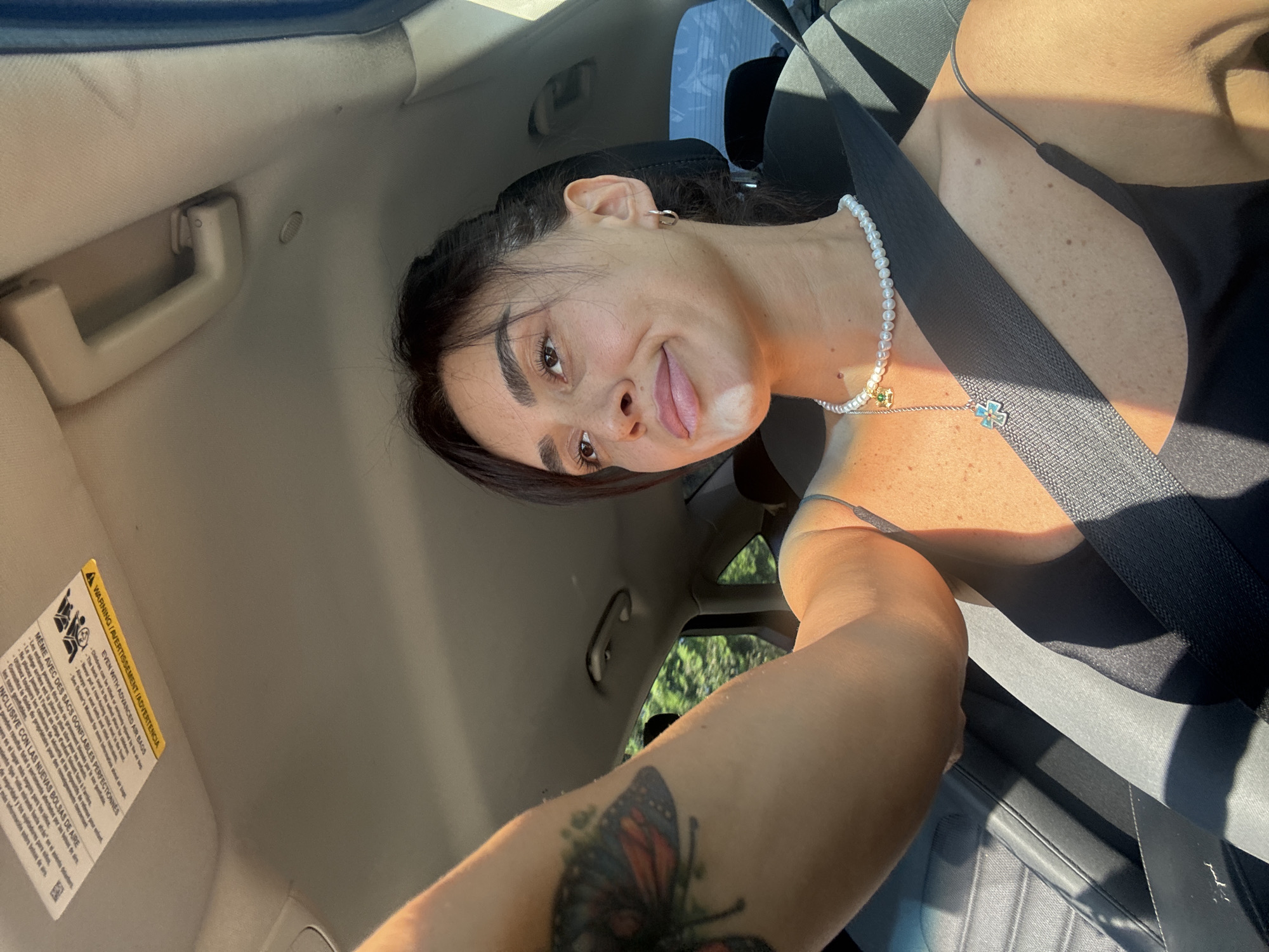 A woman is sitting in the passenger seat of a car, taking a selfie. She is wearing a black tank top, a pearl necklace with a blue and green pendant, and a silver hoop earring. A portion of a butterfly tattoo is visible on her left arm. Sunlight is casting shadows across her face and body. The car's interior is visible, including the dashboard, sun visor, and seatbelt. The scene appears to be taking place during the daytime, likely in a residential area given the trees visible outside the car window. There is no indication in the image that this is in Tbilisi, Georgia.