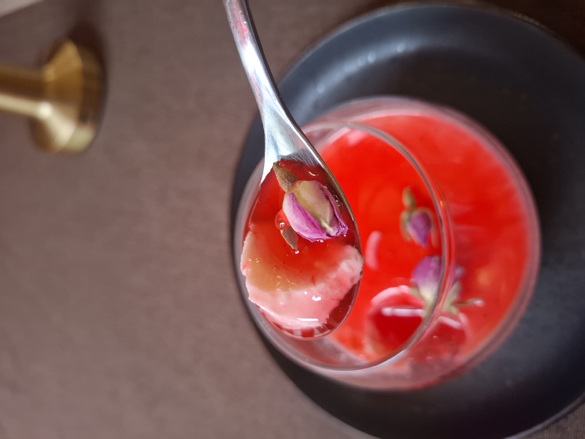 A spoon lifts a spoonful of dessert from a clear glass filled with red liquid and flower petals. The dessert appears to be a light, gelatinous substance. The glass sits on a dark gray plate. The background consists of a brown table and a blurred gold-colored object. There is no indication of the location being Zadonsk, Russia.