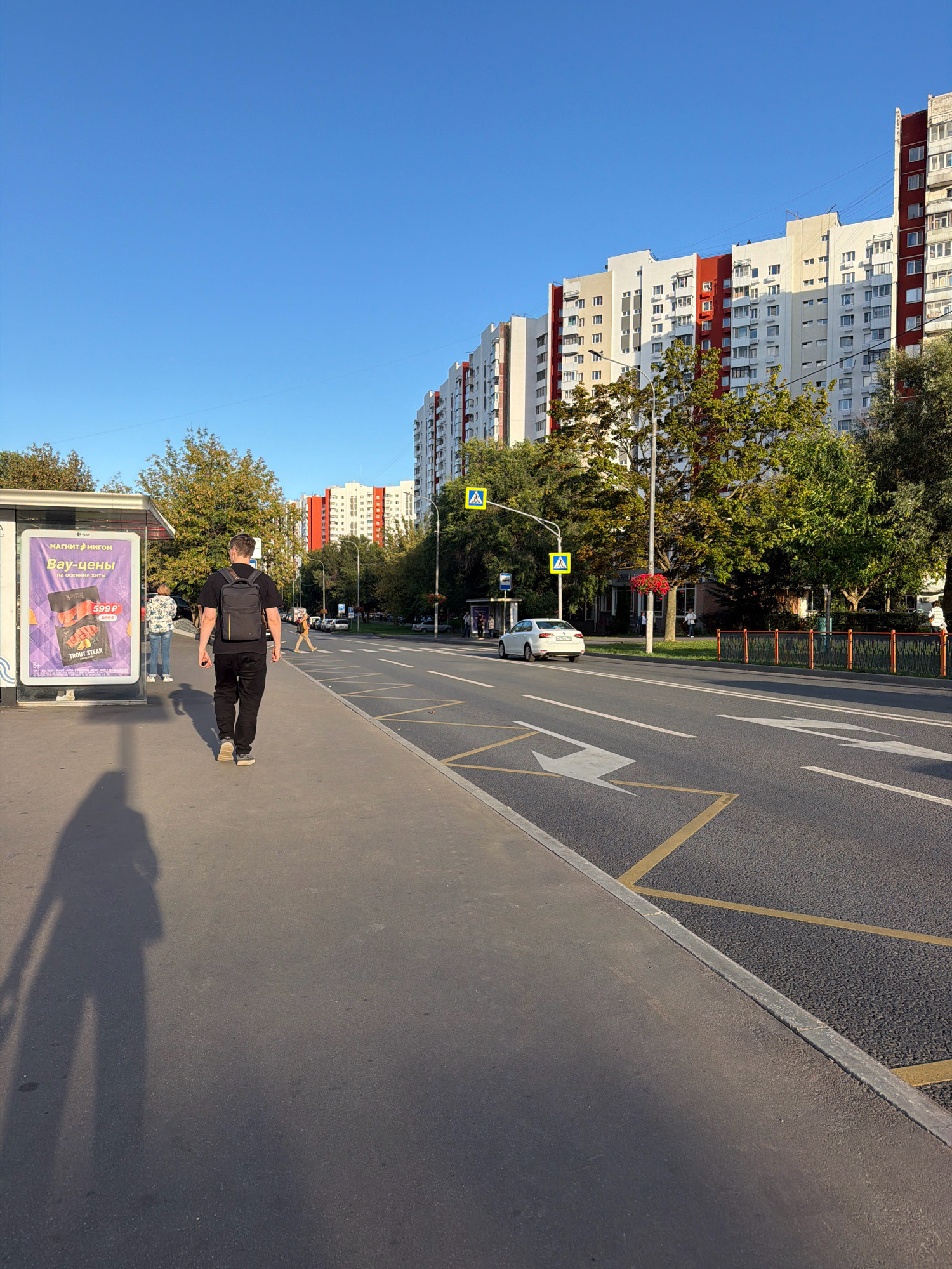 A man with a backpack walks away from the camera on a sidewalk in front of a bus stop. A large advertisement is displayed on the side of the bus stop. In the background, there are apartment buildings and trees lining a road with cars. The scene is set in Moscow, Russia, under a clear blue sky.