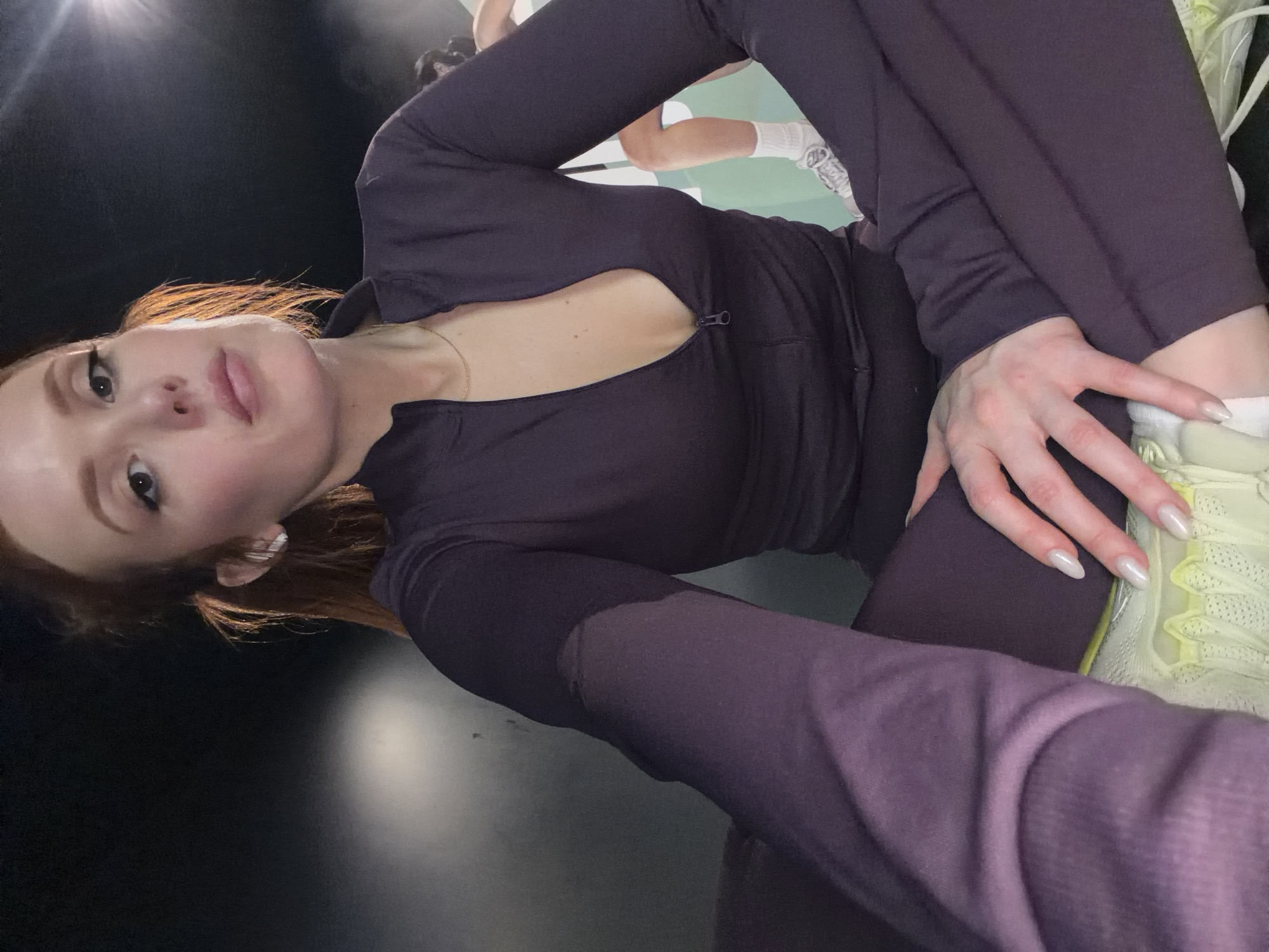 A woman is posing in a selfie, angled to the left. She has auburn hair, pale skin, and is wearing a dark purple long-sleeve top with a small zipper opening and matching pants. Her left hand is resting on her knee. There is another person visible in the background, wearing white socks and shoes. The setting appears to be indoors, with a dark backdrop. It is not possible to determine the specific location in Paris from the information available in the image.