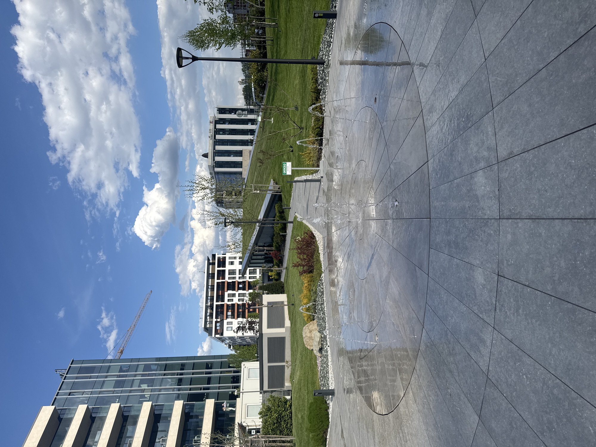 This image depicts a modern urban landscape in Sofia, Bulgaria, under a clear blue sky with scattered white cumulus clouds, suggesting a sunny day, likely mid-morning or afternoon. No people are visible in the scene.
In the foreground, a grey tiled pedestrian area features an active ground-level water fountain, with several jets of water spraying upwards from circular patterns integrated into the paving. The tiles appear wet from the water. Adjacent to this, a meticulously maintained green lawn stretches across the midground, dotted with young trees and decorative shrubs, some displaying reddish foliage. A large, light-colored rock is placed within a gravel bed that separates the paved path from the grass. Dark, slender street lamps are intermittently spaced throughout the green areas.
The background is dominated by modern architecture. To the left, a tall building with a glass and light-colored concrete facade is partially visible, with a construction crane extending into the sky behind it, indicating ongoing development. Towards the center and right, two contemporary buildings with distinct white and dark-striped facades, featuring multiple windows and balconies, rise above the landscape. Between the foreground elements and the background buildings, several modular structures, possibly temporary offices or utility units, are situated within the green spaces. A small green sign, clearly displaying the text "OFFICEO," is visible in the grass. The setting suggests a newly developed commercial or residential area with emphasis on modern design and public green spaces.