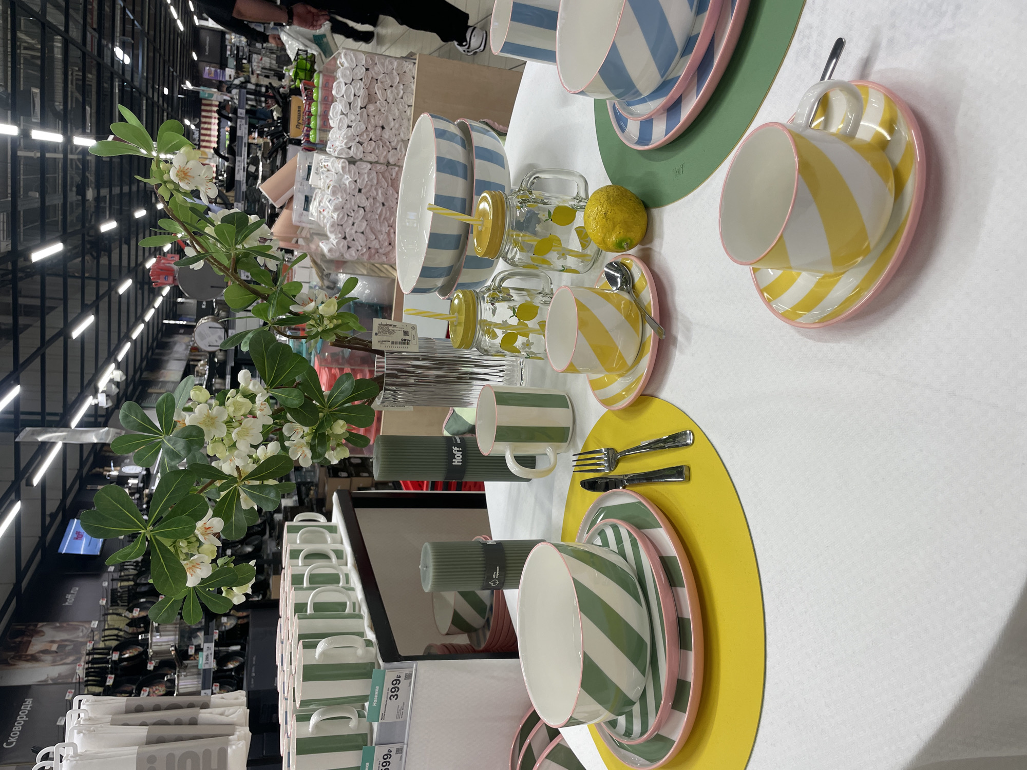 The image shows a display of tableware in a store, possibly a department store, showcasing different color schemes. The foreground features a white tablecloth arranged with place settings. On the left, there are dishes and mugs with green and white stripes, plates with pink rims, green placemats, a fork, and a knife. To the right are yellow and white striped mugs and saucers with pink rims, accompanied by glasses with lemon patterns and yellow straws, and blue and white striped bowls and plates. A single lemon is placed near the blue tableware. In the background, shelves are stocked with various merchandise, indicating that the scene is set inside a retail environment. A floral arrangement with white flowers and green leaves adds a decorative touch to the display. Tags and labels on some of the products suggest price information and possibly brand names. The store is likely located in Sochi, Russia, based on Cyrillic text seen in the background. The image focuses on a curated presentation of kitchenware, emphasizing color coordination and modern design.