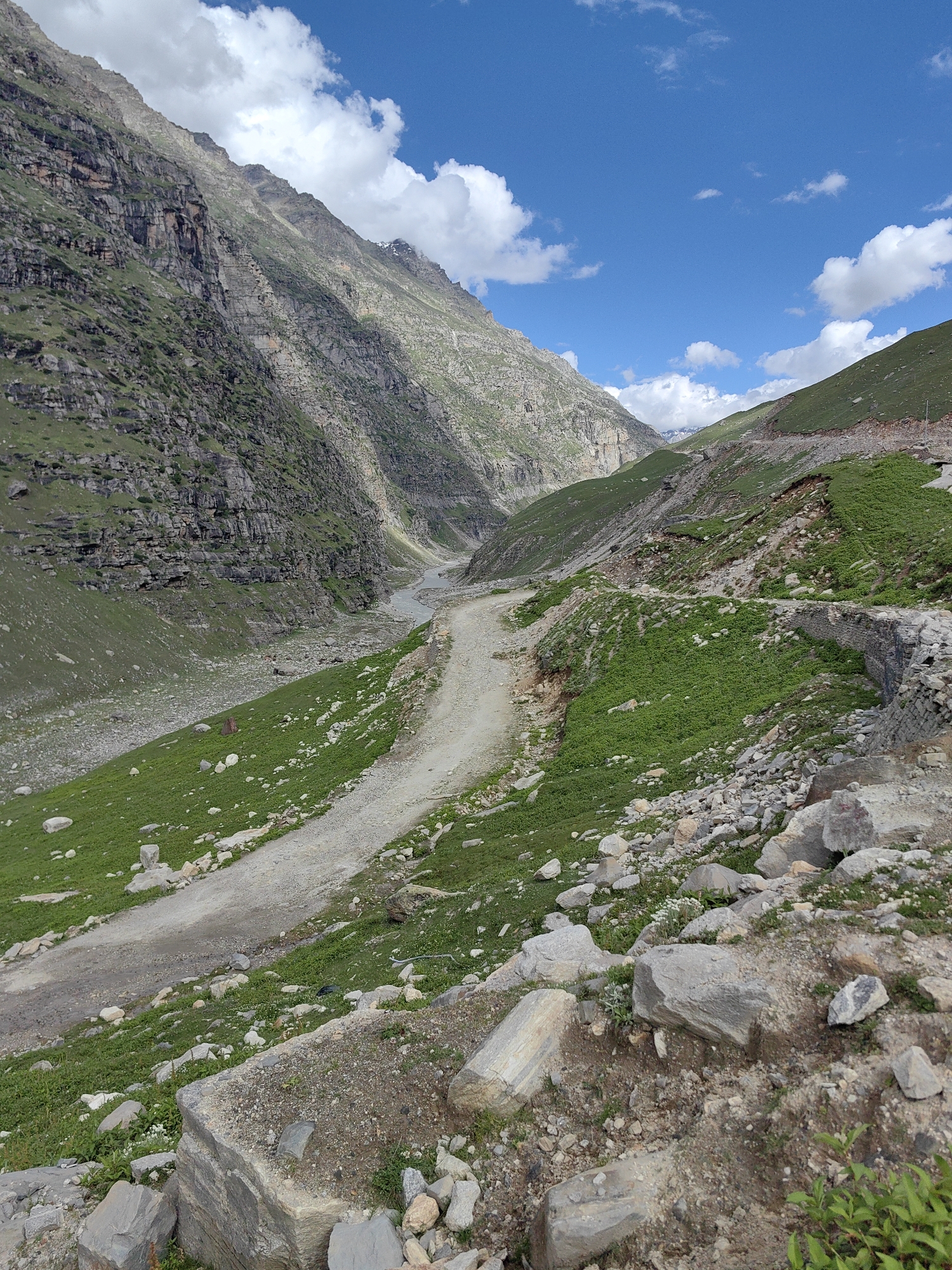 An eye-level, vertical, full shot captures a rugged mountain landscape under a bright blue sky dotted with fluffy white clouds. A narrow, unpaved road winds its way up a steep, grassy slope, bordered by a low stone wall on the right. The road is the central focus, leading the eye into the distance where a valley opens up, revealing a river and more mountains. The terrain is characterized by rocky outcrops, patches of green grass, and steep, layered rock faces on the left. The scene appears to be a remote mountain pass in India.