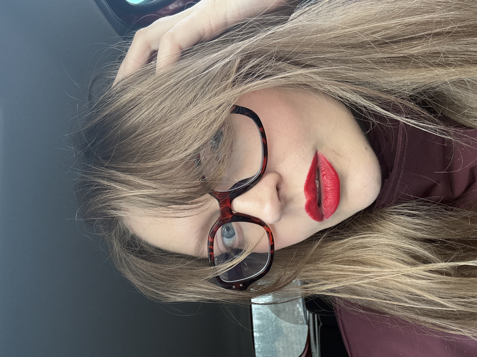 A woman is positioned at an angle, wearing red lipstick and tortoise-shell glasses. Her hand is in her hair. The background is a dark gray color. There is nothing in the image to indicate the location is Izhevsk, Russia.