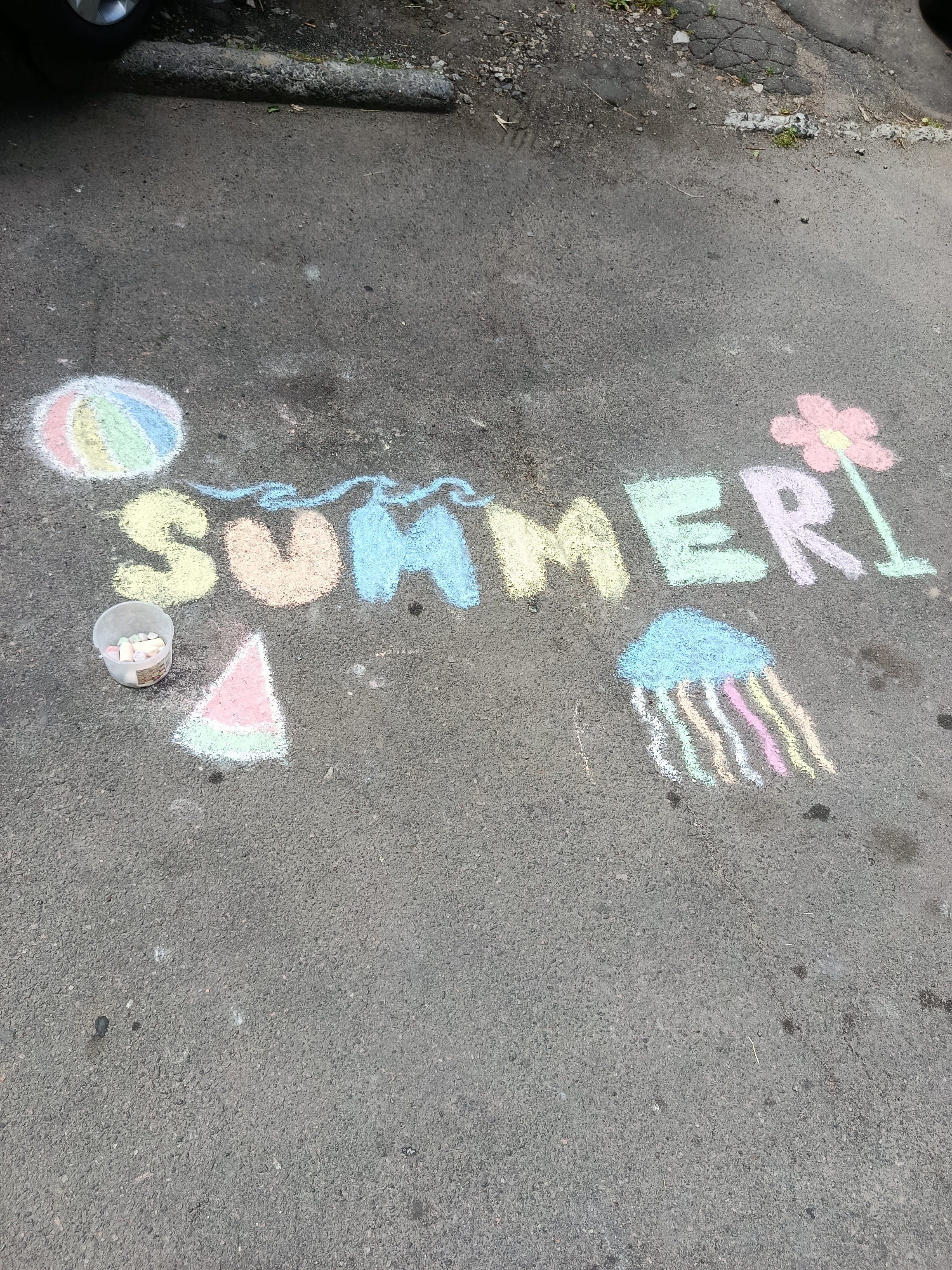The image shows the word "SUMMER" written in colorful chalk on asphalt. To the left of the word, there is a drawing of a beach ball and a small container of chalk pieces. Below the "SUM" part of the word, there is a drawing of a slice of watermelon. To the right of the word "MER," there is a drawing of a pink flower and a blue jellyfish with colorful tentacles. The asphalt surface is gray and appears to be outdoors, possibly a driveway or a street. A portion of a car tire is visible at the top left of the image.