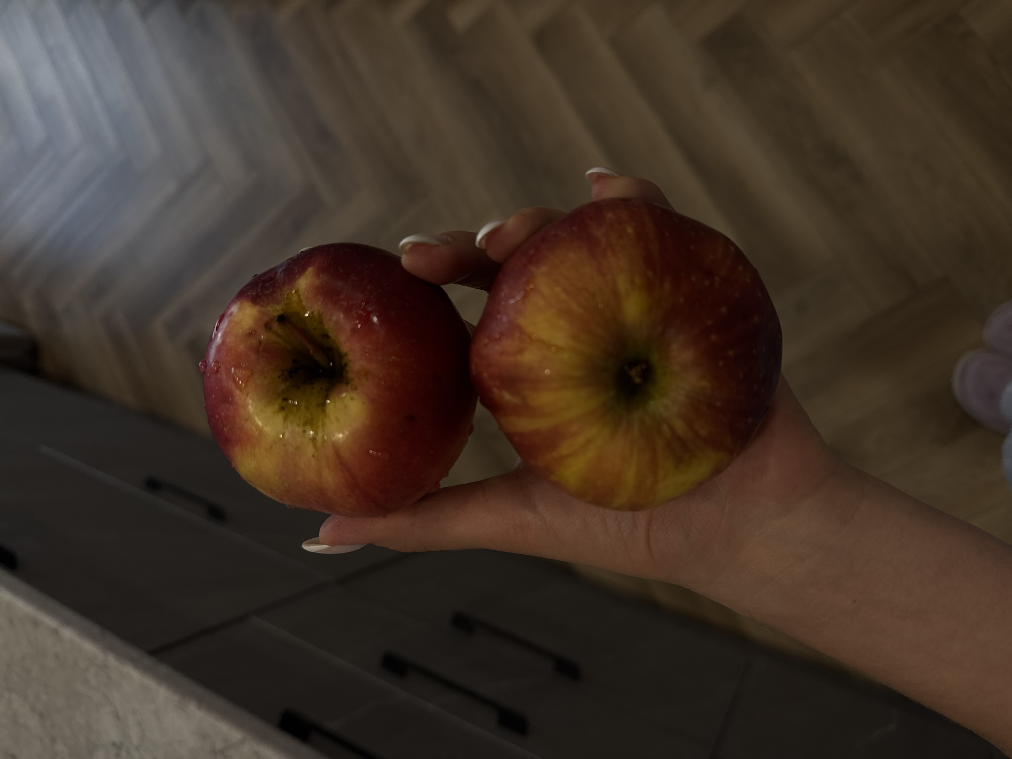 A close-up, slightly overhead shot captures a human hand holding two ripe apples against a blurred indoor background. The hand, positioned centrally, is fair-skinned with visible wrinkles and texture, and features neatly manicured nails with white tips (resembling a French manicure).
The two apples, likely recently washed, are the primary focus. They are a mix of vibrant red and golden yellow hues, with visible stripes and patches of color. Both apples have numerous small water droplets clinging to their skin, indicating freshness. The stem ends of the apples are visible, showing their characteristic indentations, with the left apple's stem end facing slightly more towards the viewer.
The background is softly out of focus, providing context without distracting from the main subjects. Above the hand and apples, a wooden floor with a distinct herringbone or parquet pattern is visible, rendered in muted brown and grey tones. Below the hand, a dark countertop or cabinet surface with two indistinct dark handles can be seen. There is no visible text in the image. The lighting is soft and ambient, highlighting the texture and color of the apples and hand.