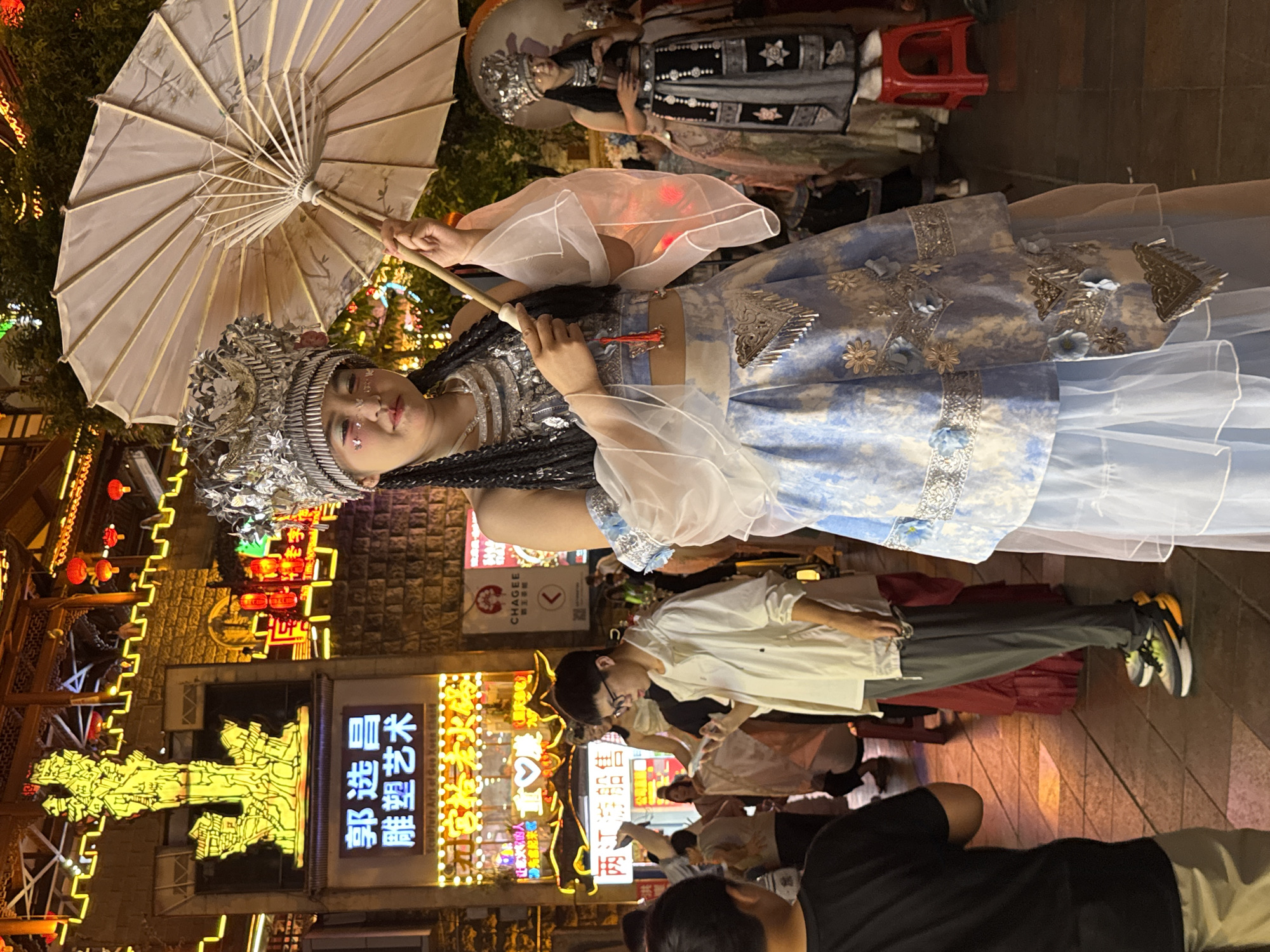 A young woman in traditional Chinese attire, adorned with a silver headdress and shimmering makeup, stands in a bustling street at night in the Yuzhong District of China. She holds a decorative umbrella, its fabric patterned with subtle imagery. Her elaborate costume features a silver ornate top, flowing translucent sleeves, and a blue and white patterned skirt with intricate silver embellishments and tassels. To her right, another woman in a similar, darker traditional outfit stands beside a red stool. In the foreground, a young man in casual attire, wearing a white jacket and grey pants, looks down at his phone. Other individuals, some in traditional dress and others in modern clothing, are visible in the background, amidst glowing neon signs and traditional Chinese architectural elements that suggest a vibrant cultural district. The scene is illuminated by artificial lights, casting a warm glow on the cobblestone street.