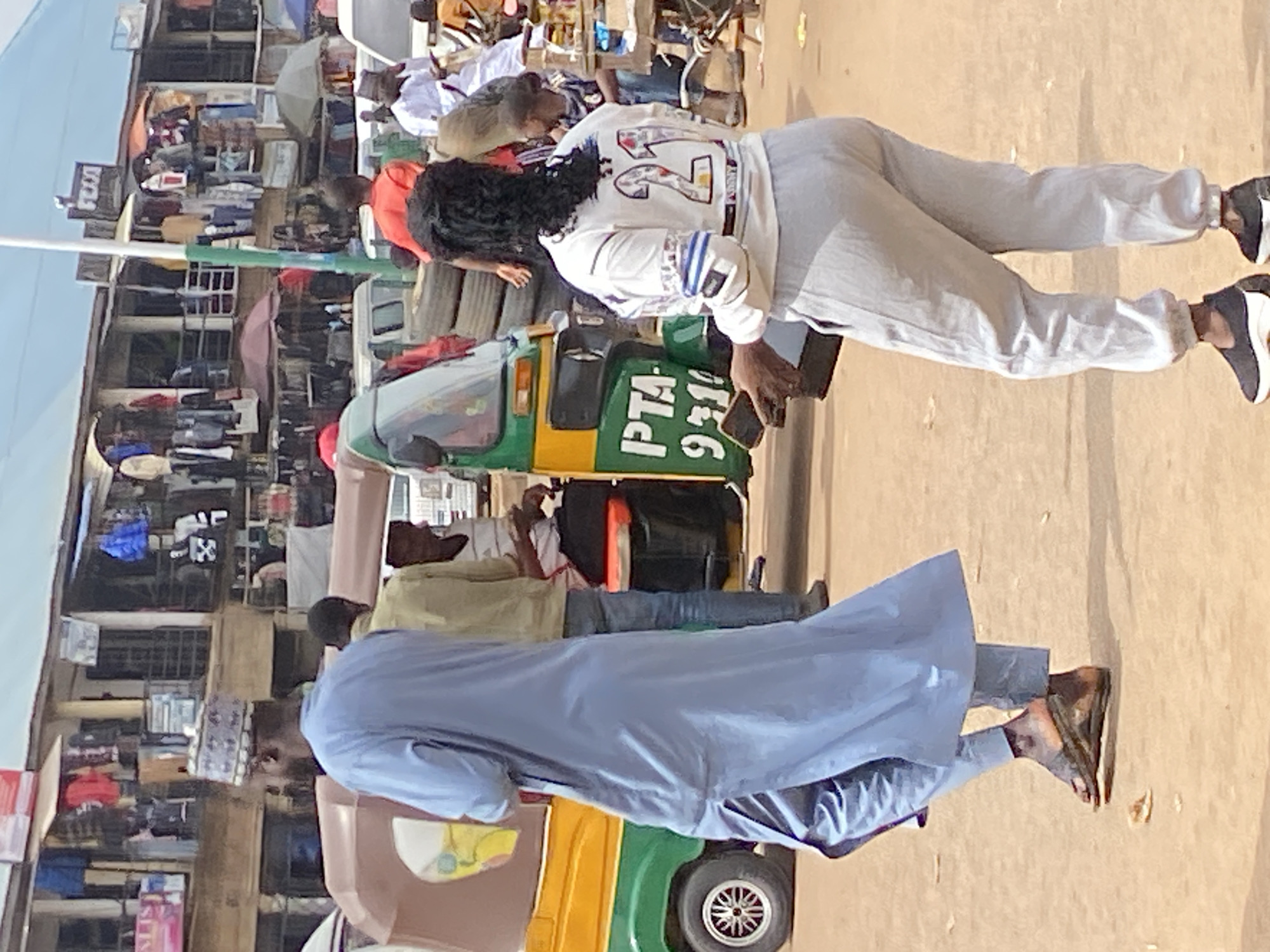 A street scene in Bukuru, Nigeria, features a bustling marketplace with stalls displaying clothing. In the foreground, a person in a light blue traditional garment and a patterned cap walks away from the camera, their feet clad in dark sandals. To their right, another person, seen from behind, wears a white long-sleeved shirt with the number "21" prominently displayed on the back, and light gray sweatpants. This individual holds a dark rectangular object, possibly a phone, in their right hand. Behind them, a colorful auto-rickshaw, green and yellow, is parked. Other individuals are visible in the background, including one in a yellow shirt and another in a white outfit. The ground is dusty, and market structures with awnings and hanging clothes are visible in the background.