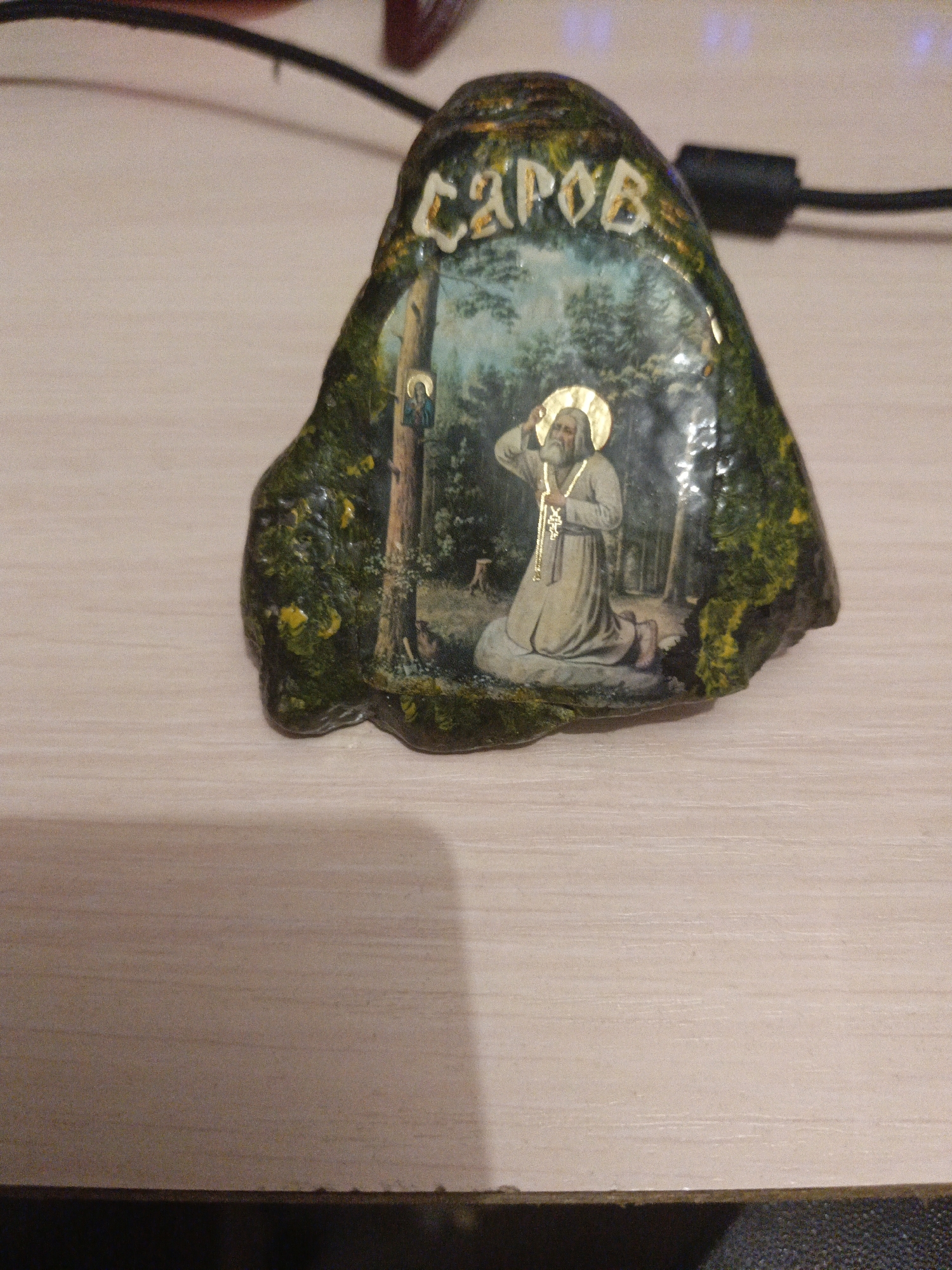 A decorative stone with a painted religious scene and text sits on a wooden surface. The stone is irregular in shape and coated with a glossy finish, appearing green and textured with yellow accents, possibly representing moss or foliage. The painted scene depicts a man with a halo, kneeling in a forest setting, holding a key. A small icon is affixed to a tree on the left side of the scene. Above the painted image, white letters spell out "CAPOB." The background of the image shows a wooden table with a dark cable running across it. The context of the city of Uren, Russia is not visually discernible from the image itself.