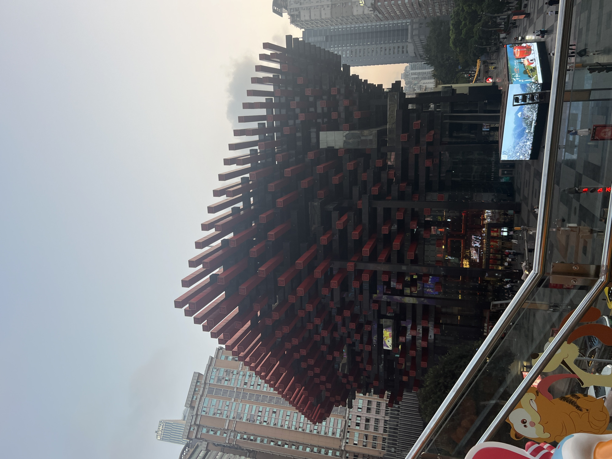 The image shows the Raffles City Chongqing skyscraper in Yuzhong District, China. It depicts an unusual structure with protruding rectangular beams in red and black against a backdrop of other tall buildings. The sky appears overcast. In the foreground, there is a glimpse of a modern glass railing, with cartoon decorations visible below the railing. Two large electronic screens are also visible to the right, showing different scenes. People can be seen walking in the area between the skyscraper and the camera's position.